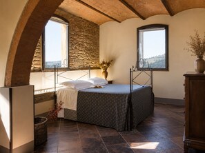 Un dormitorio con cama, dos ventanas, pared de piedra y vistas al campo.