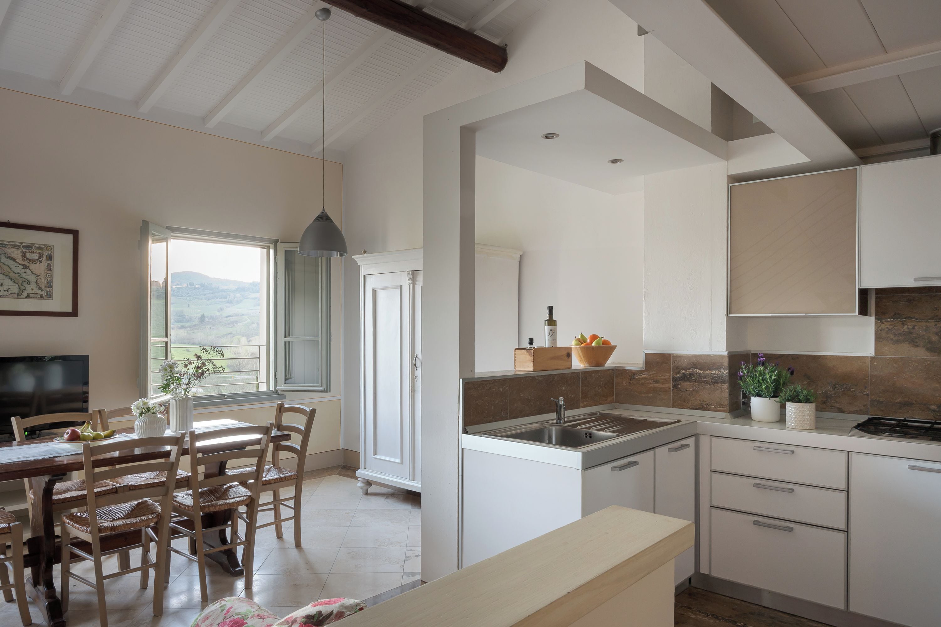 Una cocina moderna con armarios blancos, un comedor con sillas de madera y vistas al campo a través de un amplio ventanal.