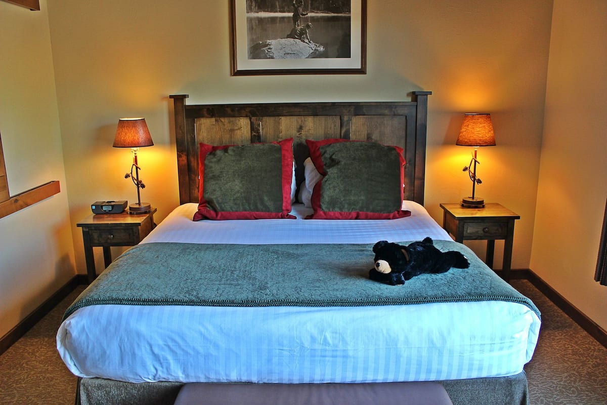 A hotel room with a bed, two bedside tables, a lamp on each table, a framed picture above the bed, and a teddy bear on the bed.