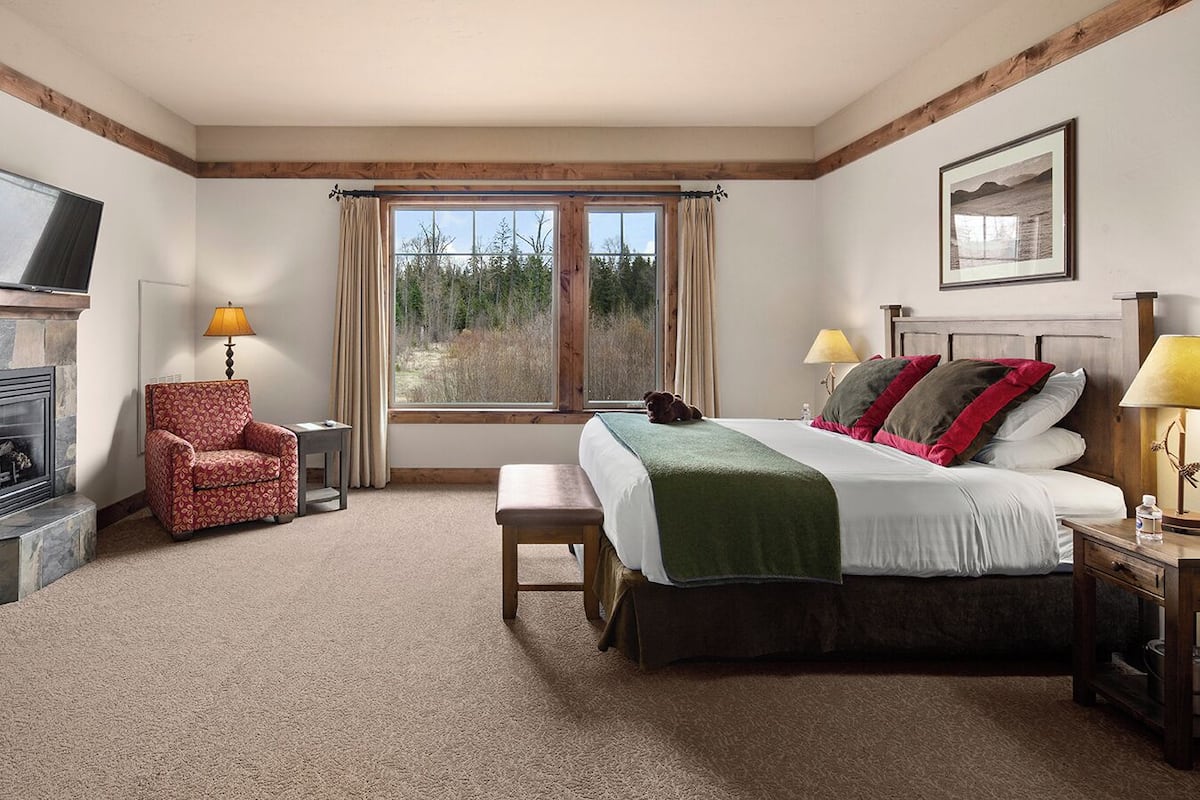 A bedroom with a large bed, a chair, a small table, a lamp, a fireplace, and a window with curtains.