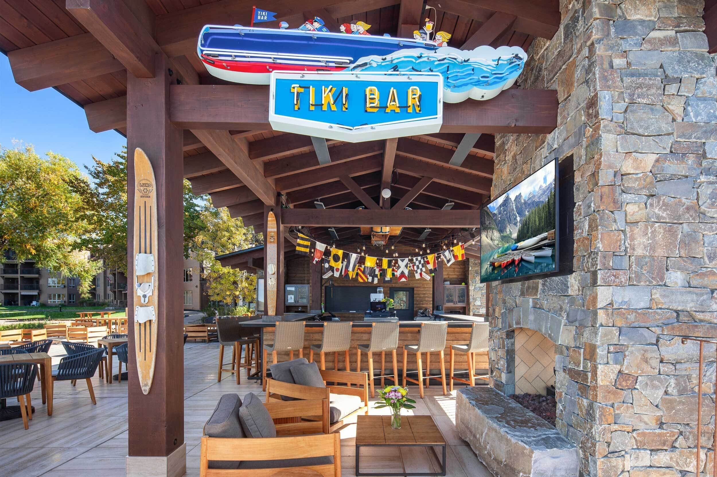 Un bar tiki a l'aire lliure amb mobiliari de fusta, una paret de pedra i un televisor penjat.