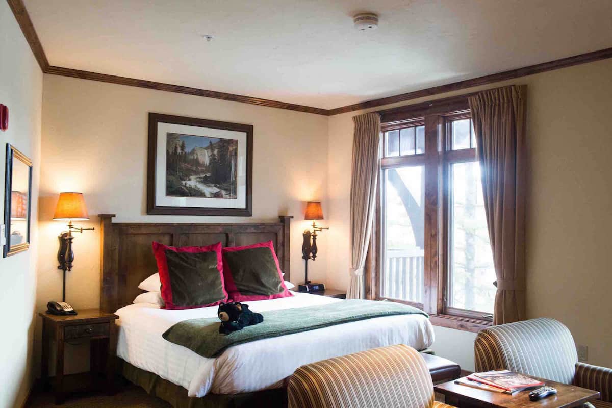A hotel room with a large bed, two bedside lamps, a painting above the bed, a window with curtains, a chair, and a table with a book and remote control.
