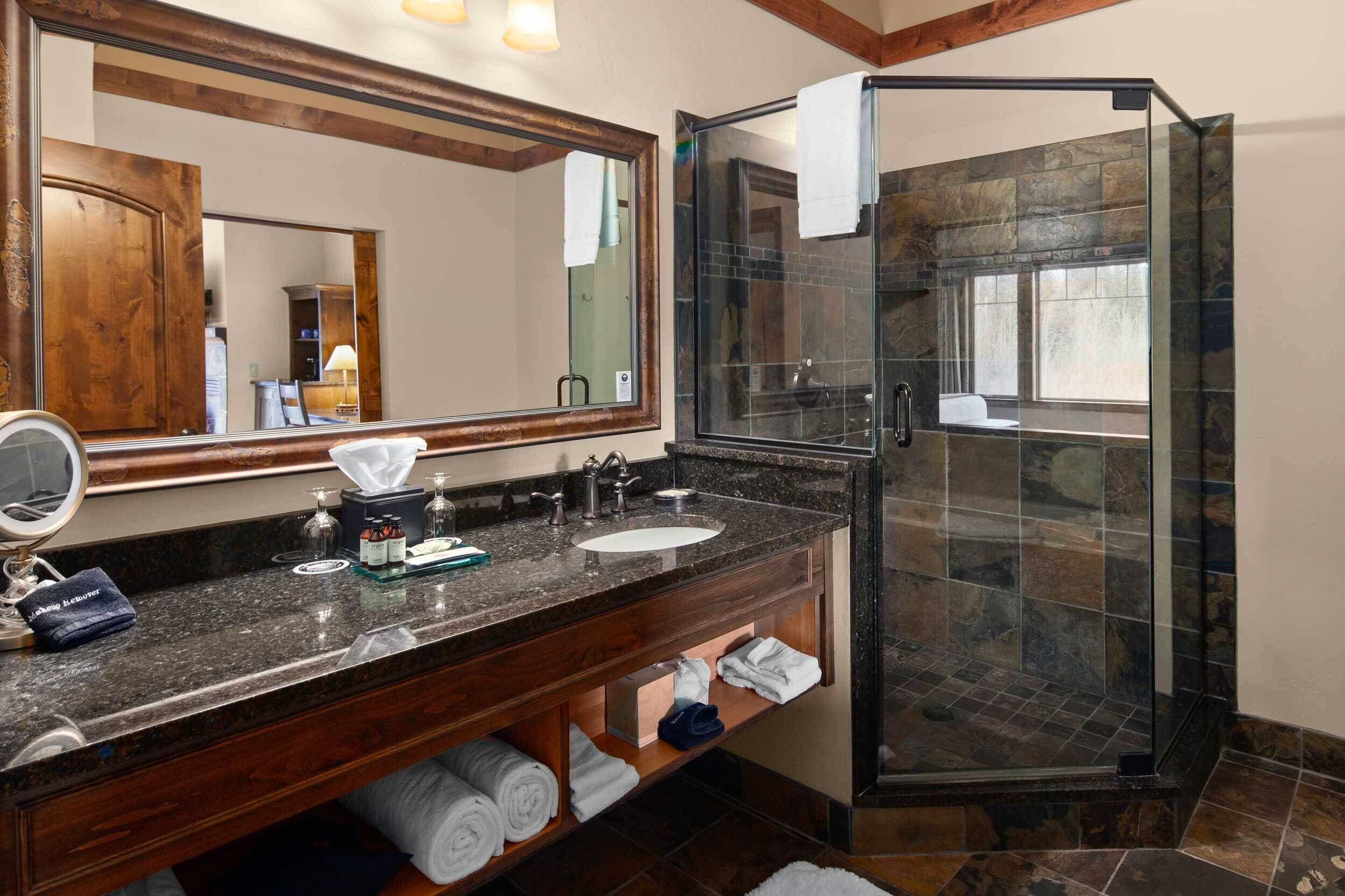 Un baño con encimera de granito, un espejo grande, cabina de ducha con mampara de vidrio y tocador de madera.