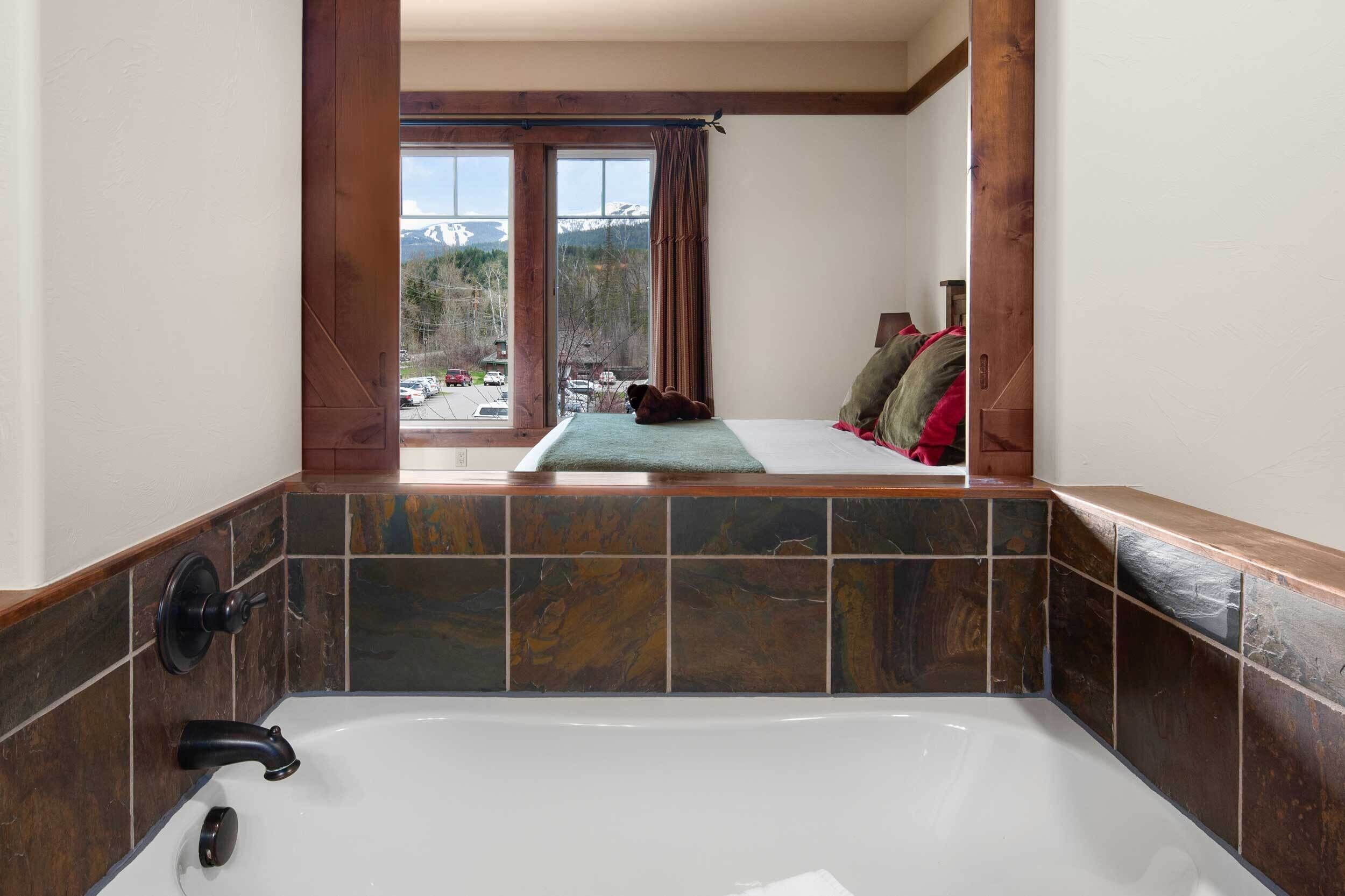 A bathroom with a large bathtub, a window with curtains, and a view of a mountain and a parking lot.