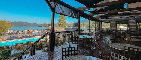An outdoor terrace with wooden flooring, tables, and chairs, overlooking a pool and a lake with mountains in the background.