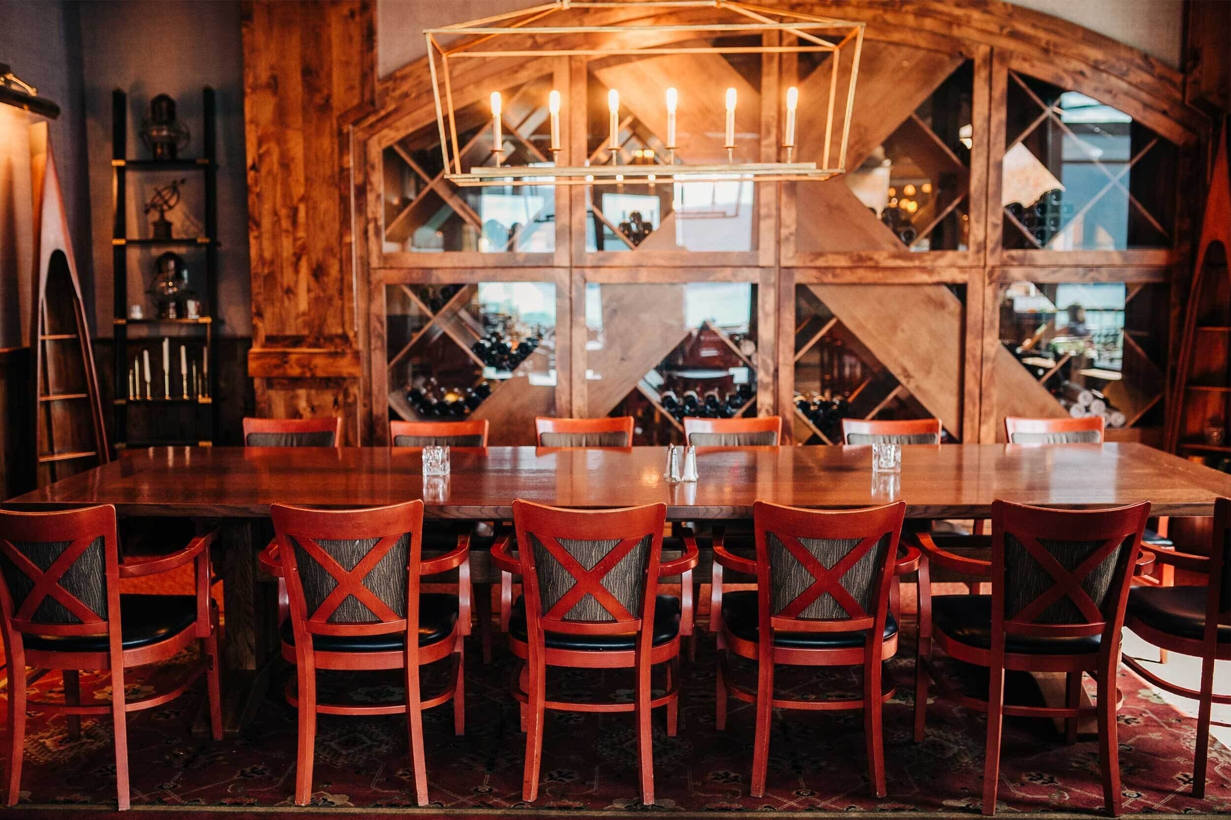 A wooden dining area with a large table and red chairs, surrounded by wooden walls and shelves with wine bottles.