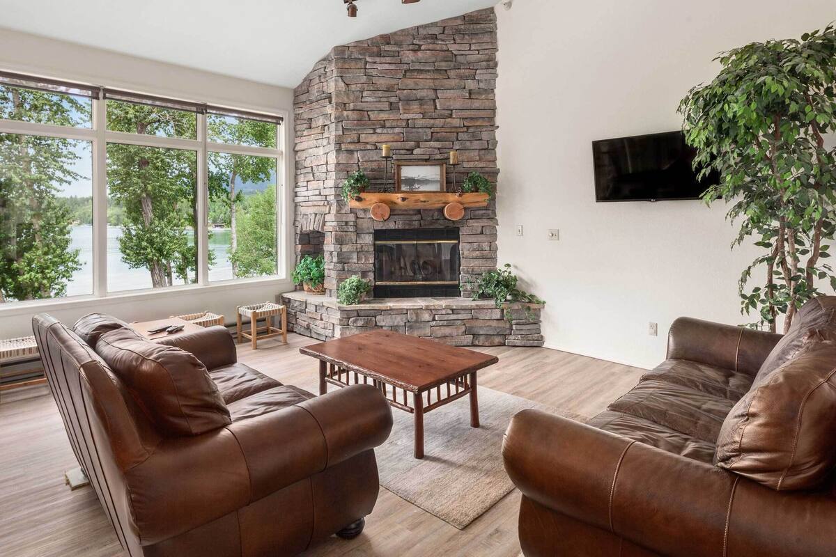 A living room with a stone fireplace, leather sofas, a wooden coffee table, and a large window with a view of trees.