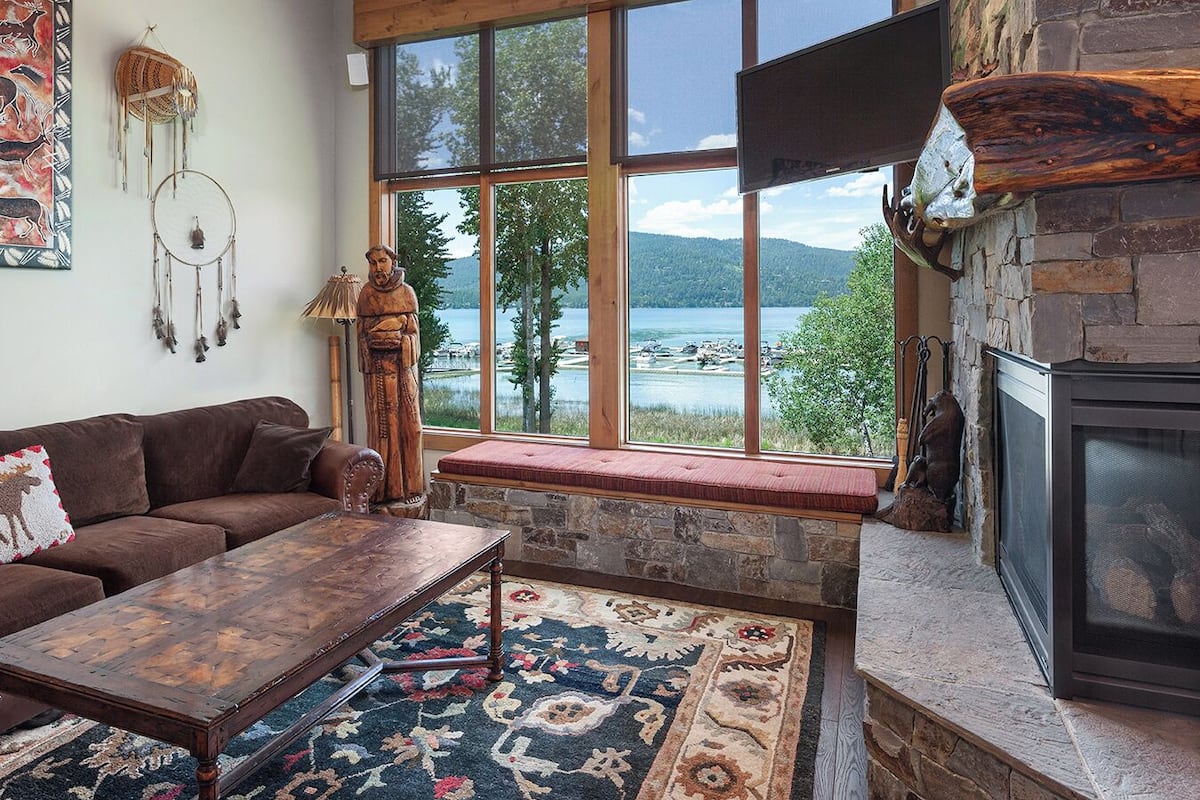 A cozy living room with a large window overlooking a lake, a stone fireplace, and rustic decor.