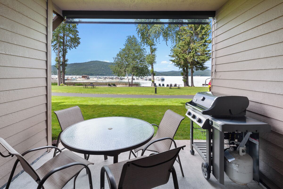 A covered patio with a grill, table, and chairs, overlooking a lake with boats and trees.