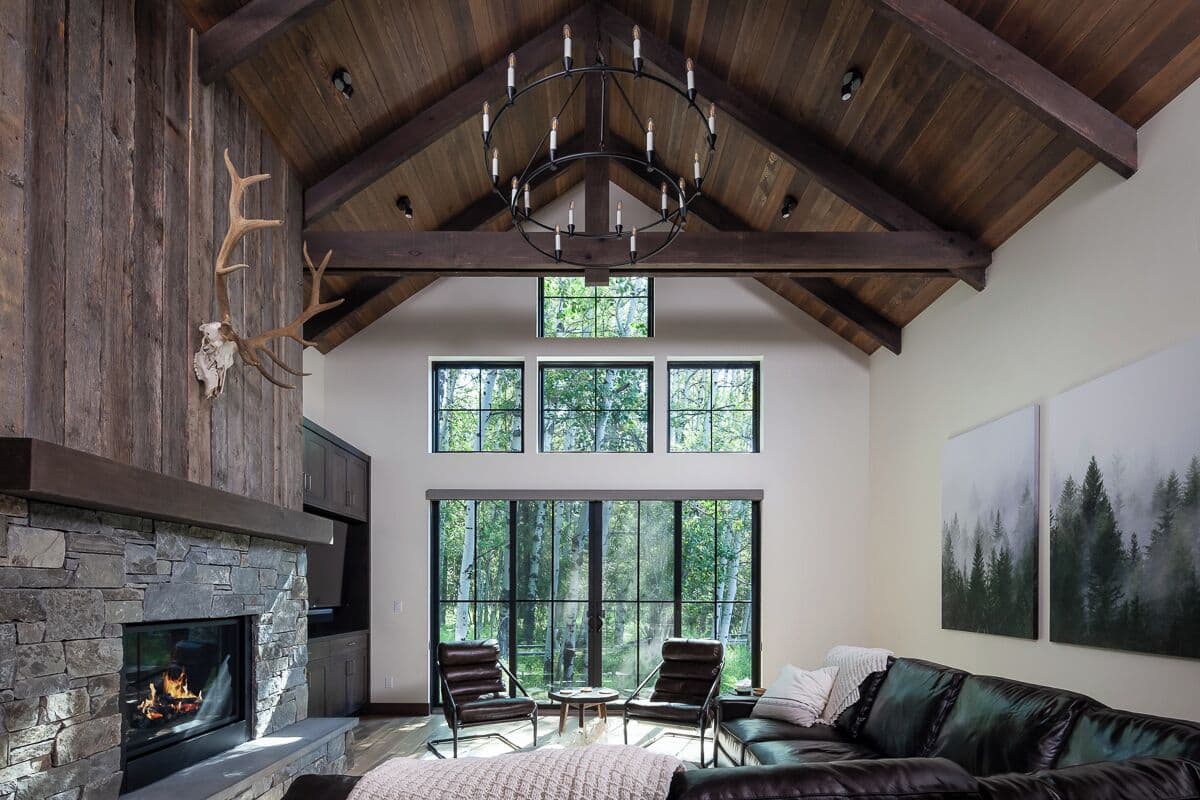A spacious living room with a stone fireplace, leather seating, large windows, and a high ceiling with exposed beams.