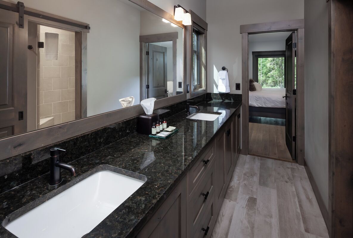 A modern bathroom with two sinks, a large mirror, and a view of a bedroom.