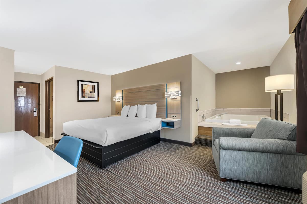 A hotel room with a bed, a desk, a chair, a sofa, a bathroom with a bathtub, and a wall-mounted mirror.