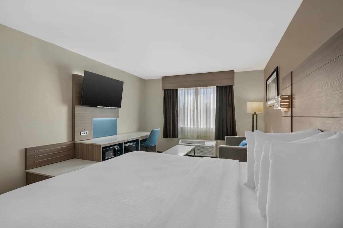 A hotel room with a large bed, a desk with a chair, a flat-screen TV mounted on the wall, a window with curtains, and a bedside table with a lamp.