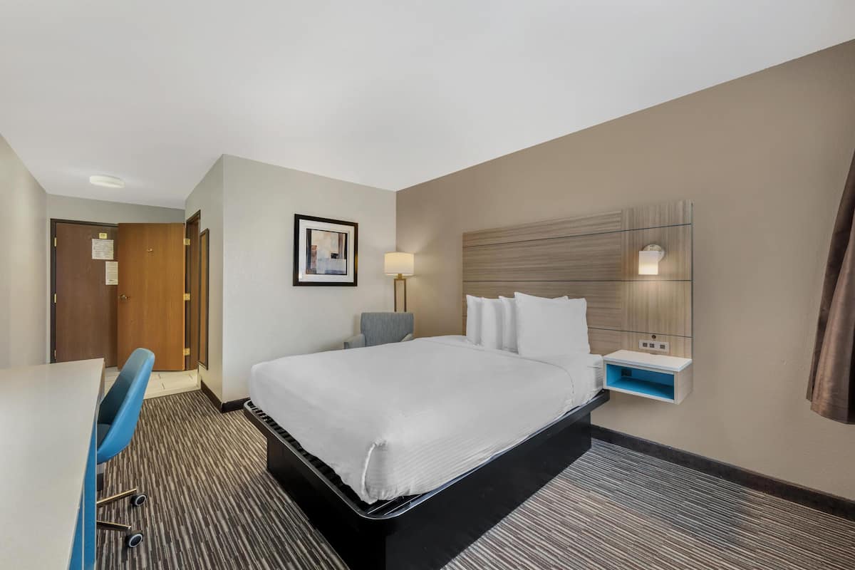 A hotel room with a bed, desk, chair, and a wall-mounted shelf with a lamp.