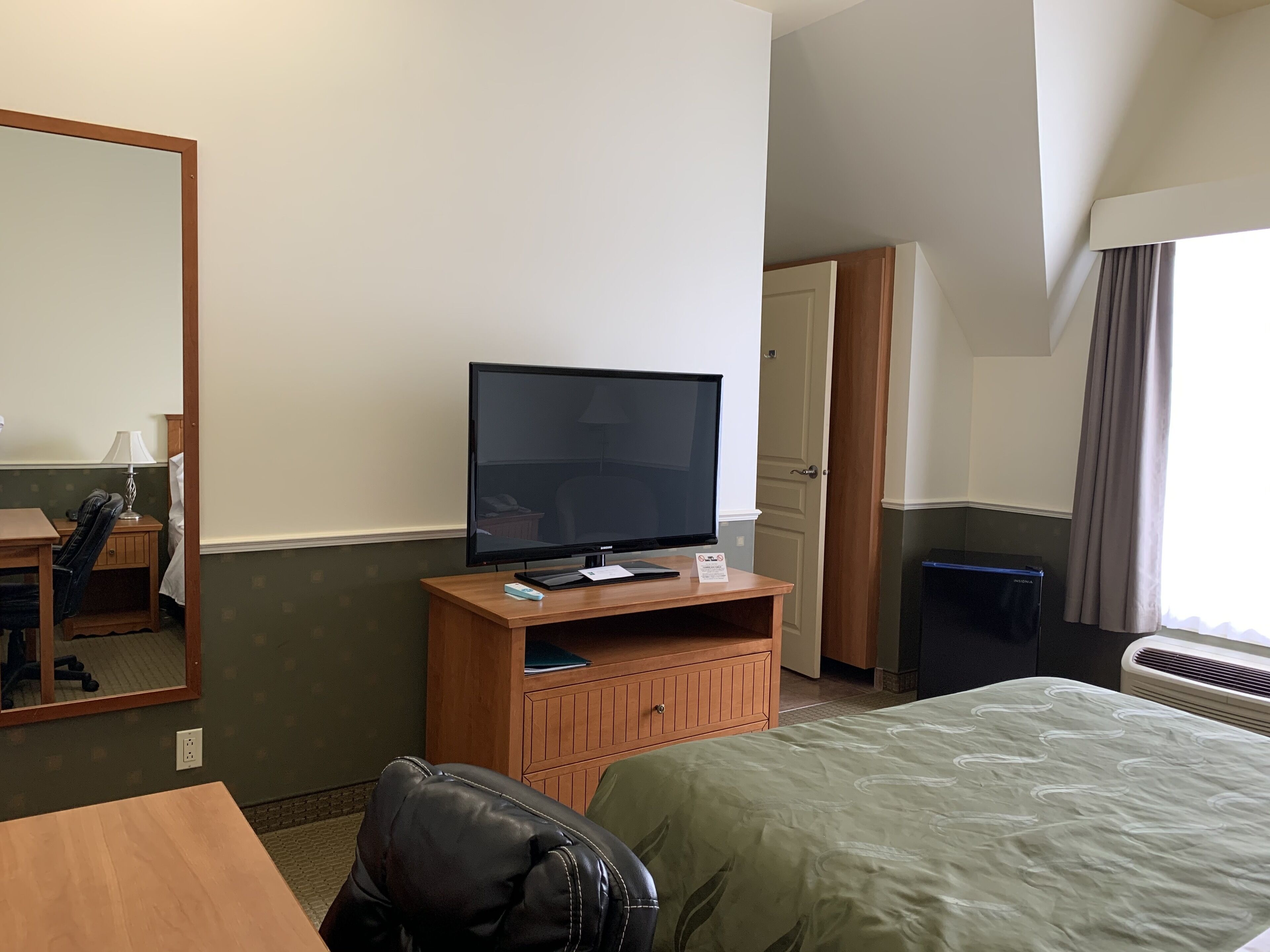 A hotel room with a bed, a desk, a chair, a television on a wooden stand, a mirror, and a window with curtains.