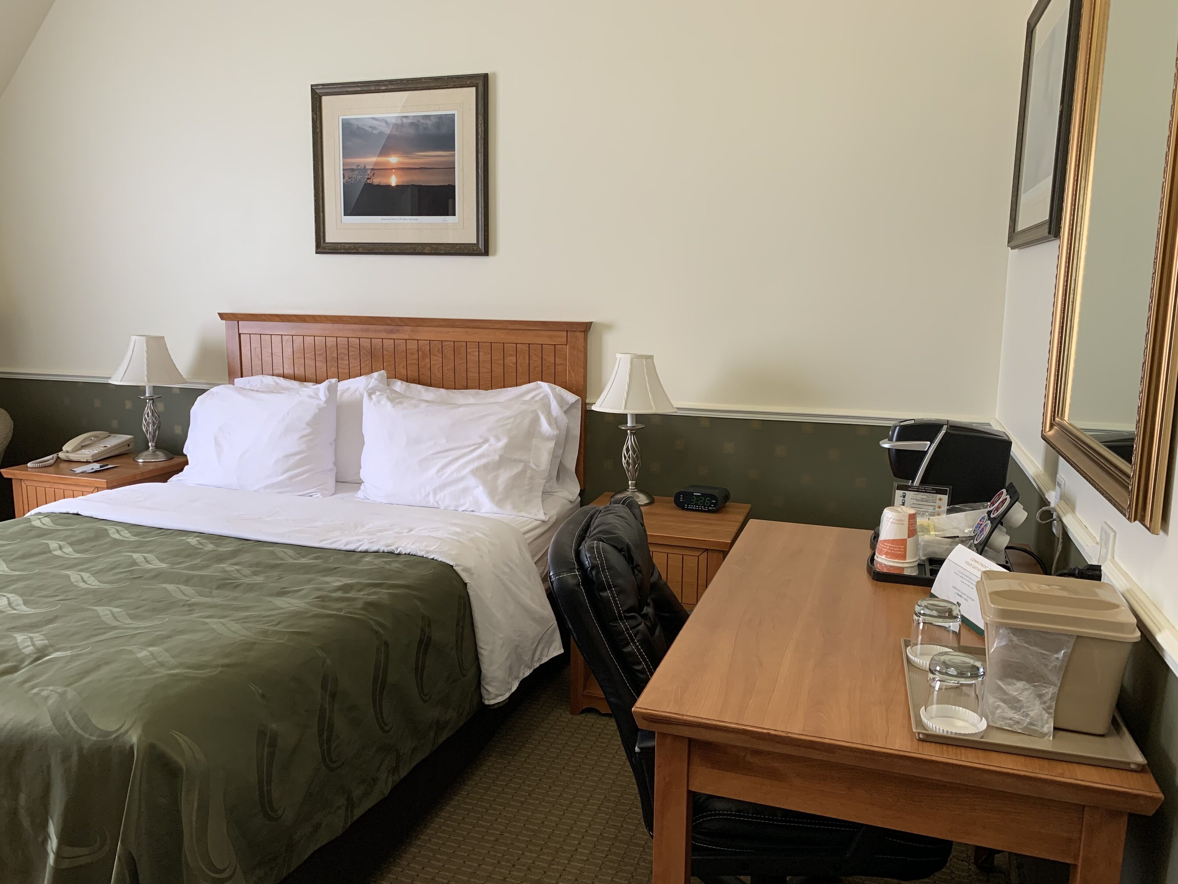 Habitación de hotel con cama, lámparas de noche, mesita de noche con teléfono, mesa con bandeja de artículos de aseo y un cuadro en la pared.