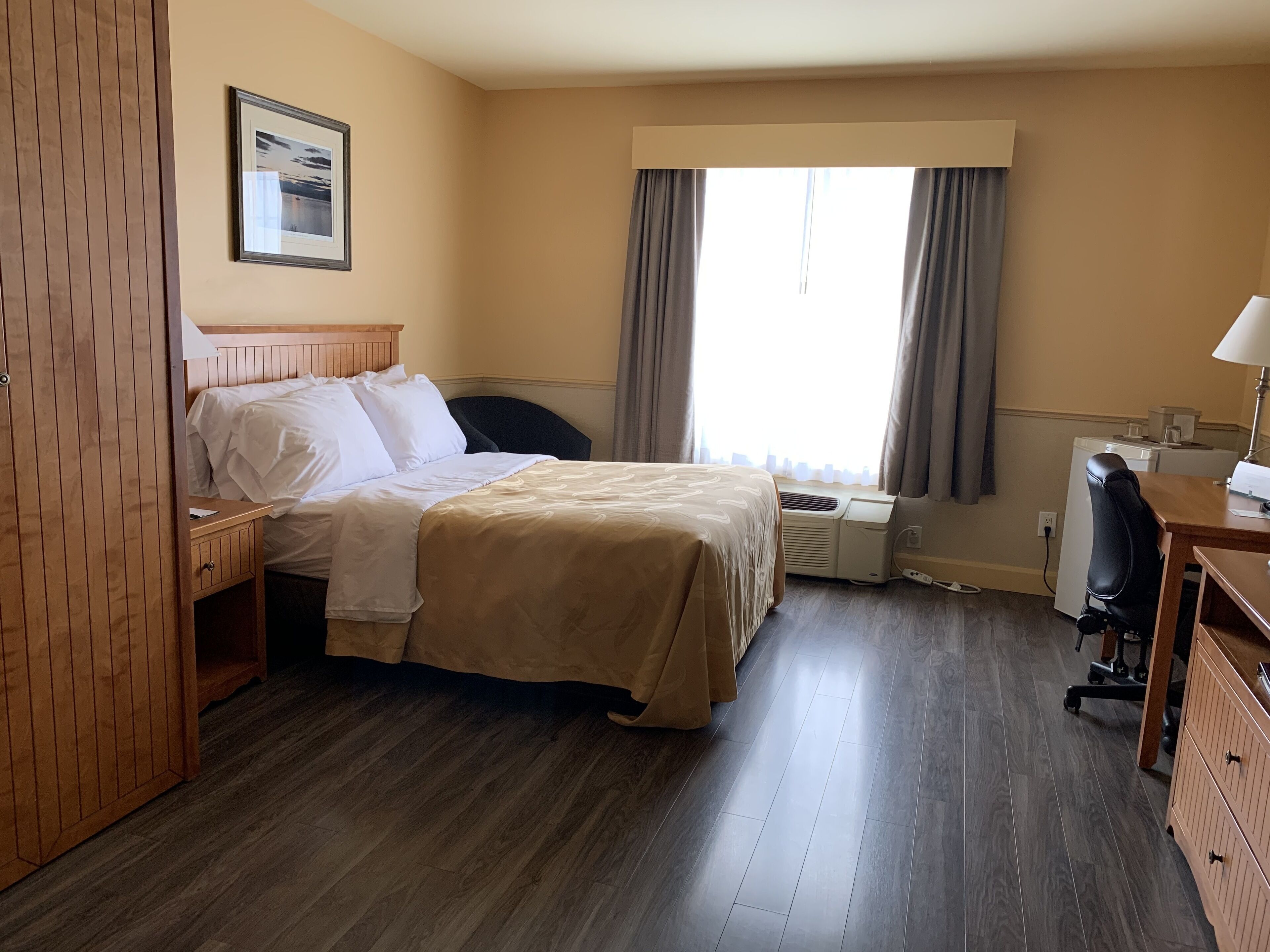 A hotel room with a bed, a desk, a chair, an air conditioning unit, and a window with curtains.