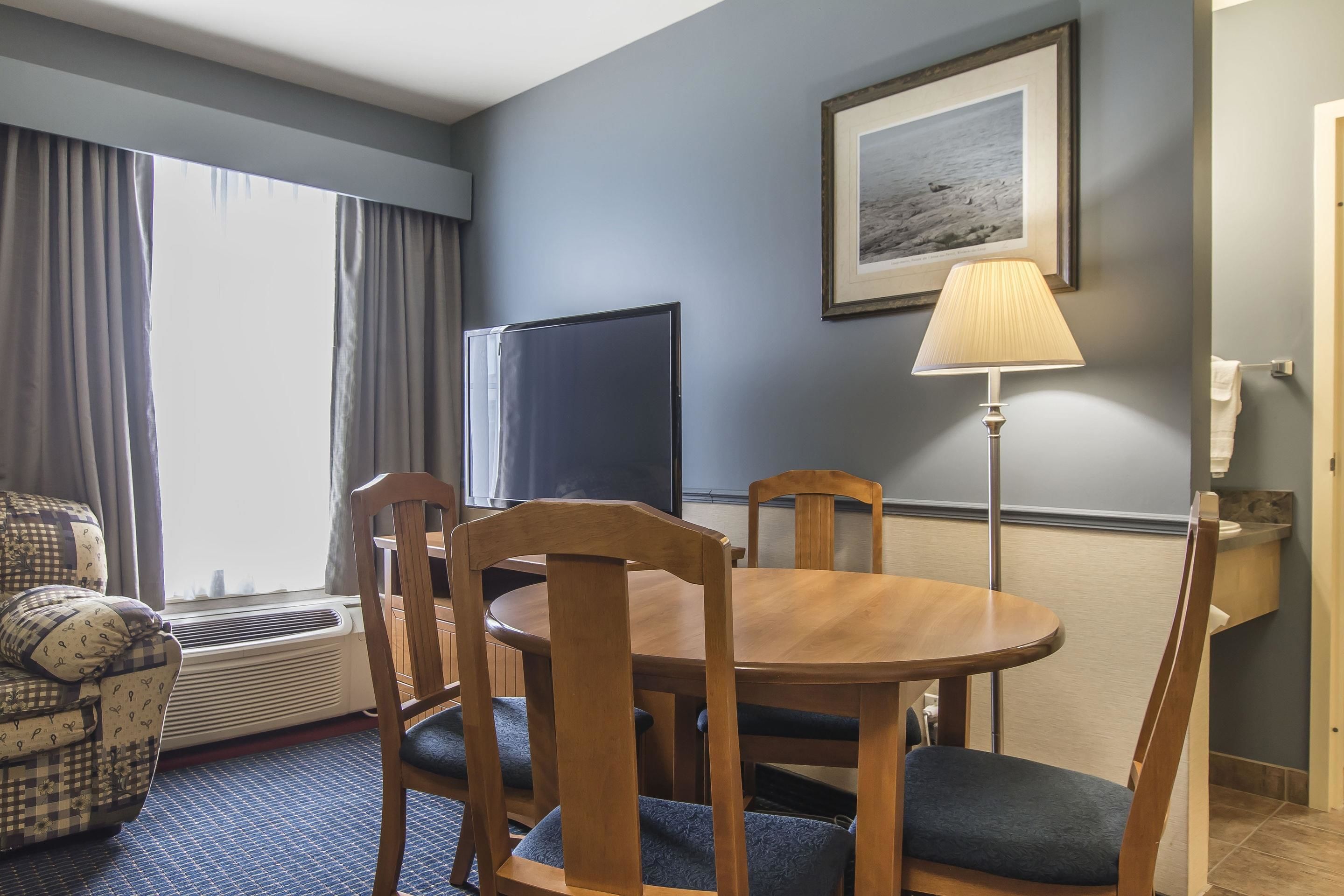 Une chambre d’hôtel dotée d’un coin repas comprenant une table ronde en bois et des chaises, un téléviseur, un tableau encadré au mur et une lampe.