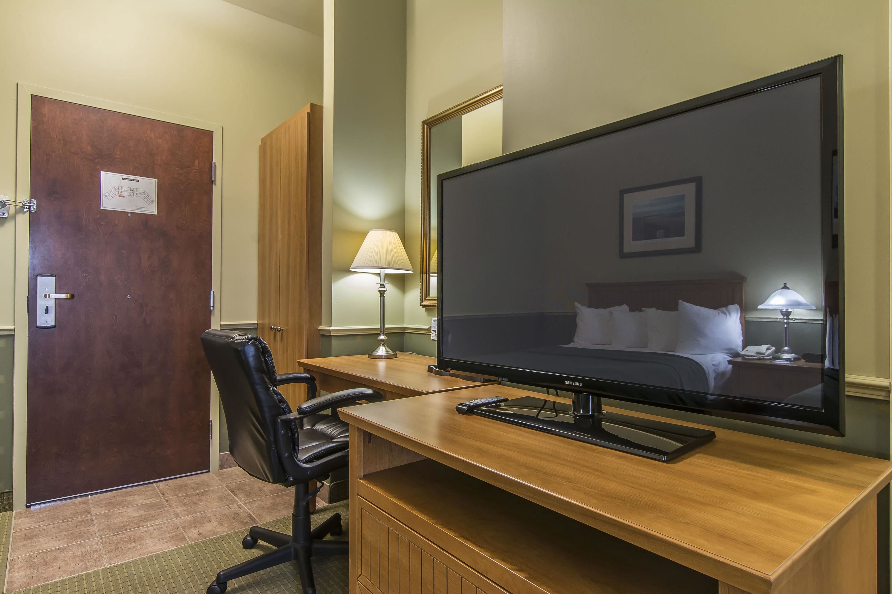 Quarto de hotel com escrivaninha de madeira, cadeira de escritório preta, televisão de tela plana, cama com lençóis brancos, mesa de cabeceira com abajur e porta com placa.