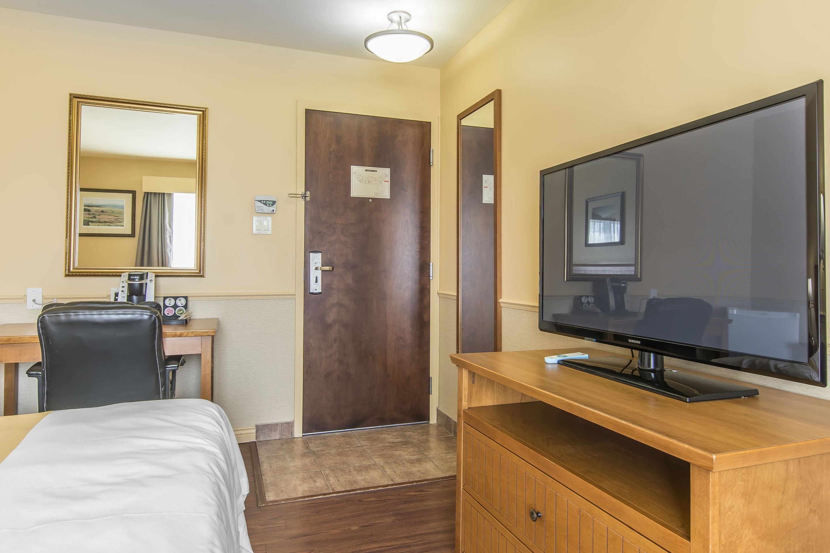 A hotel room with a bed, a desk with a chair, a television on a wooden stand, a door with a mirror, and a light fixture on the ceiling.