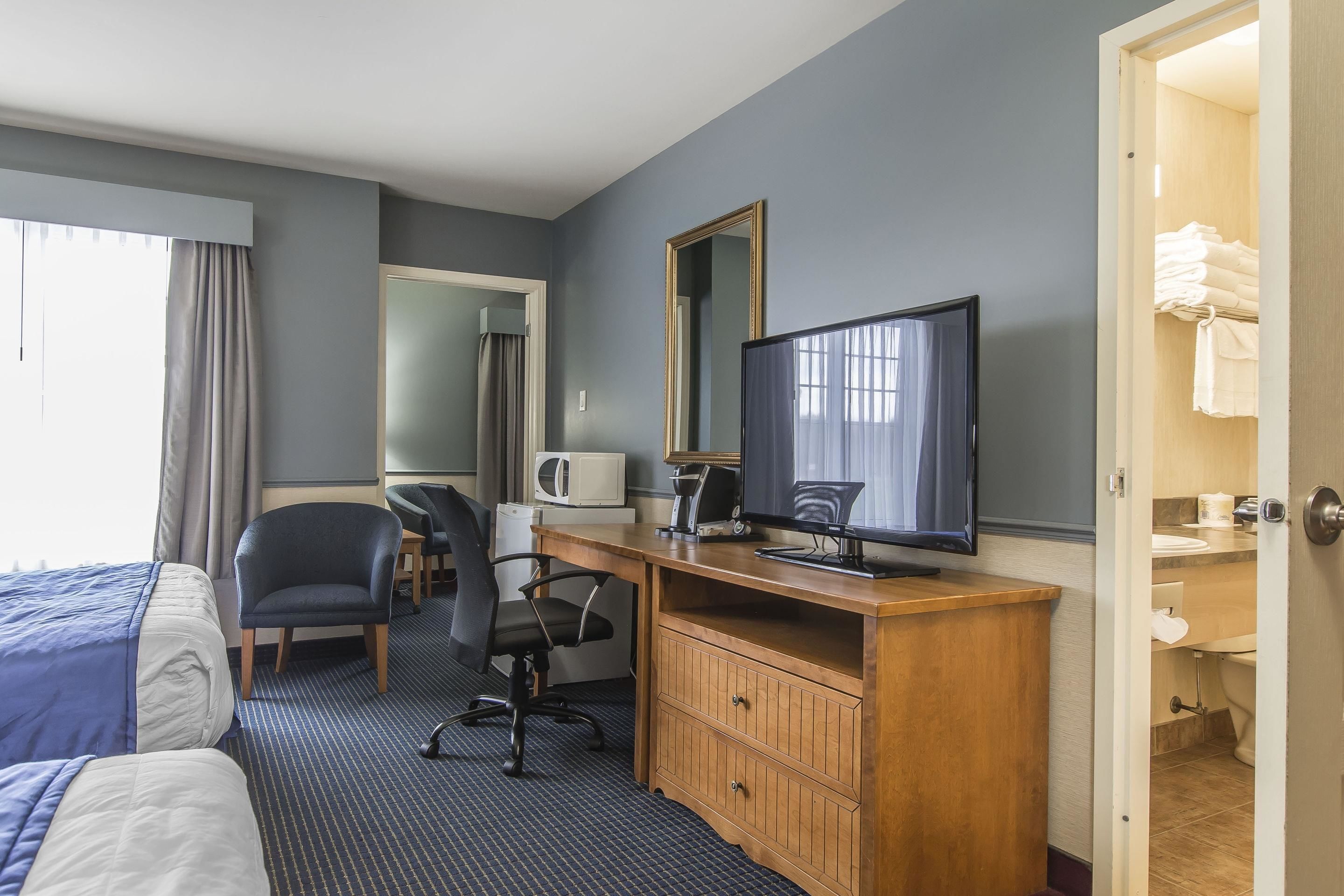 Habitación de hotel con dos camas, un escritorio con computadora, una silla, un televisor, un microondas y un baño visible a través de una puerta abierta.