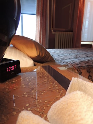 Habitación de hotel con cama, mesita de noche con reloj digital, almohada y ventana con cortinas.