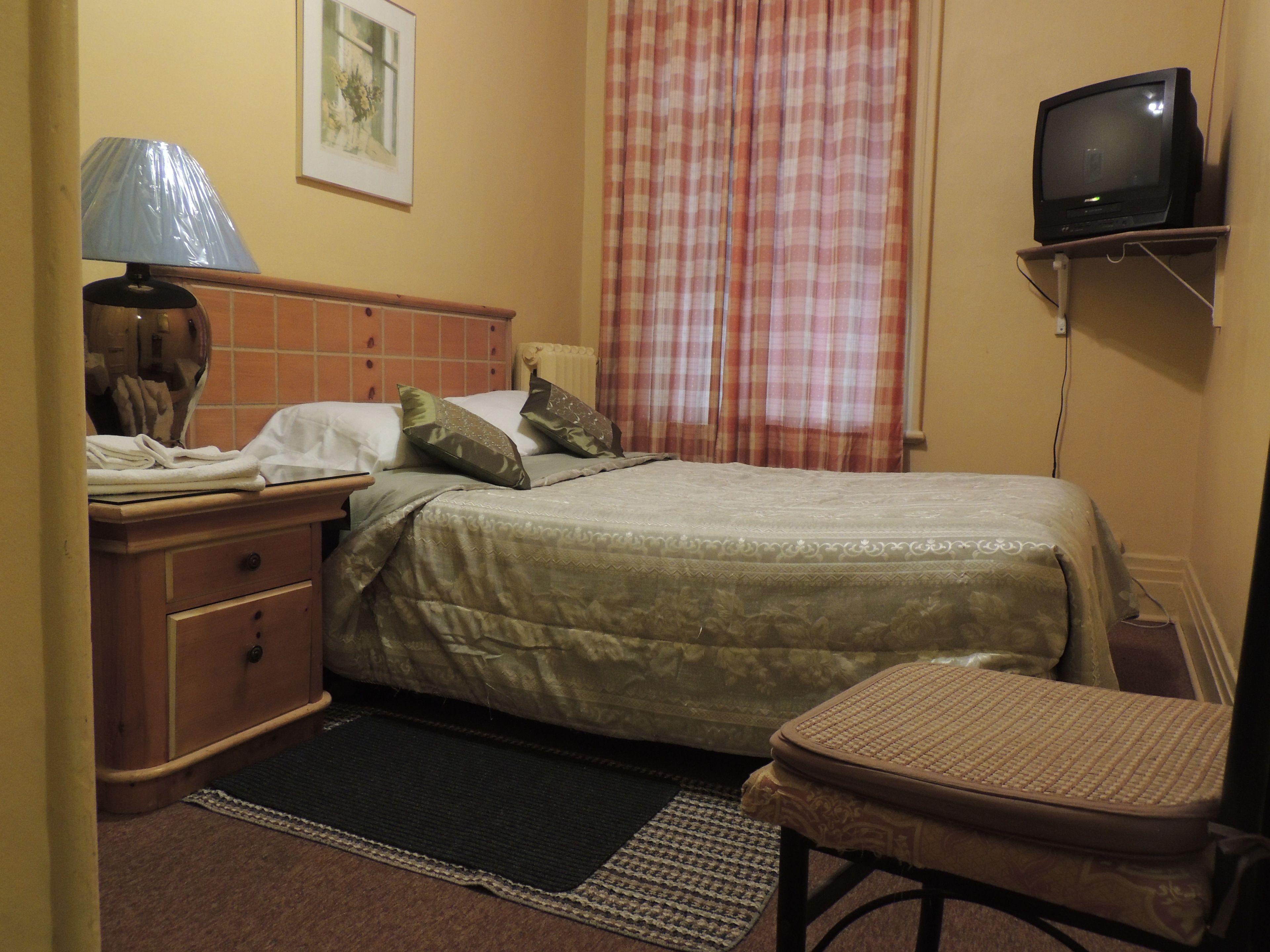 Un dormitorio con cama, mesita de noche, lámpara, silla, televisor y ventana con cortinas.