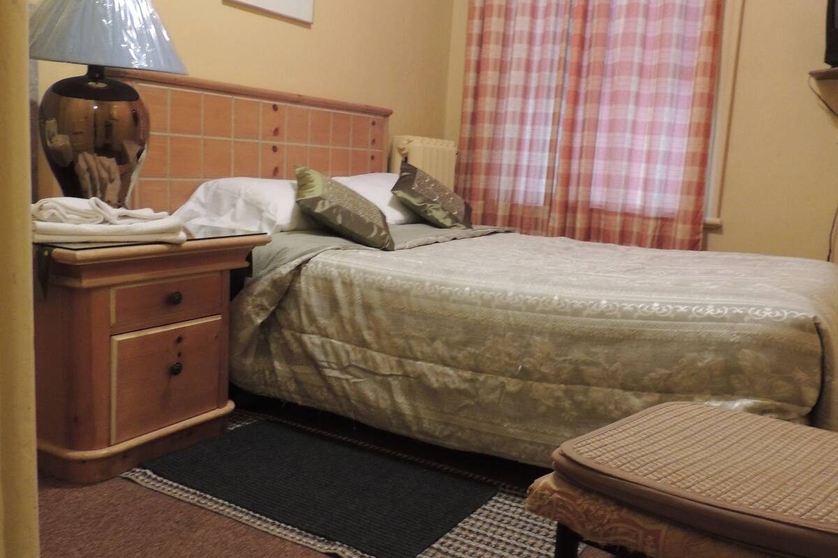 Un dormitorio con cama, mesita de noche, lámpara, sillón, radiador y ventana con cortinas.