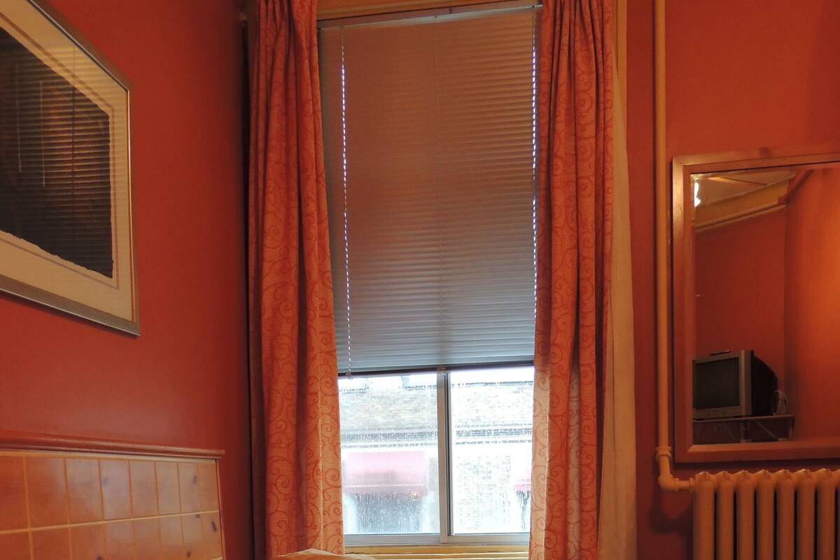 Un dormitorio con cama, almohadas, mesita de noche, radiador y ventana con cortinas.