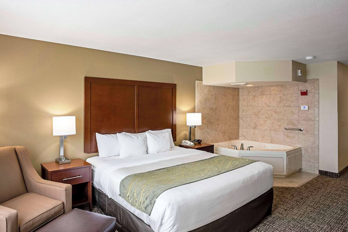 A hotel room with a large bed, a chair, a desk with a lamp, a phone, and a bathtub.