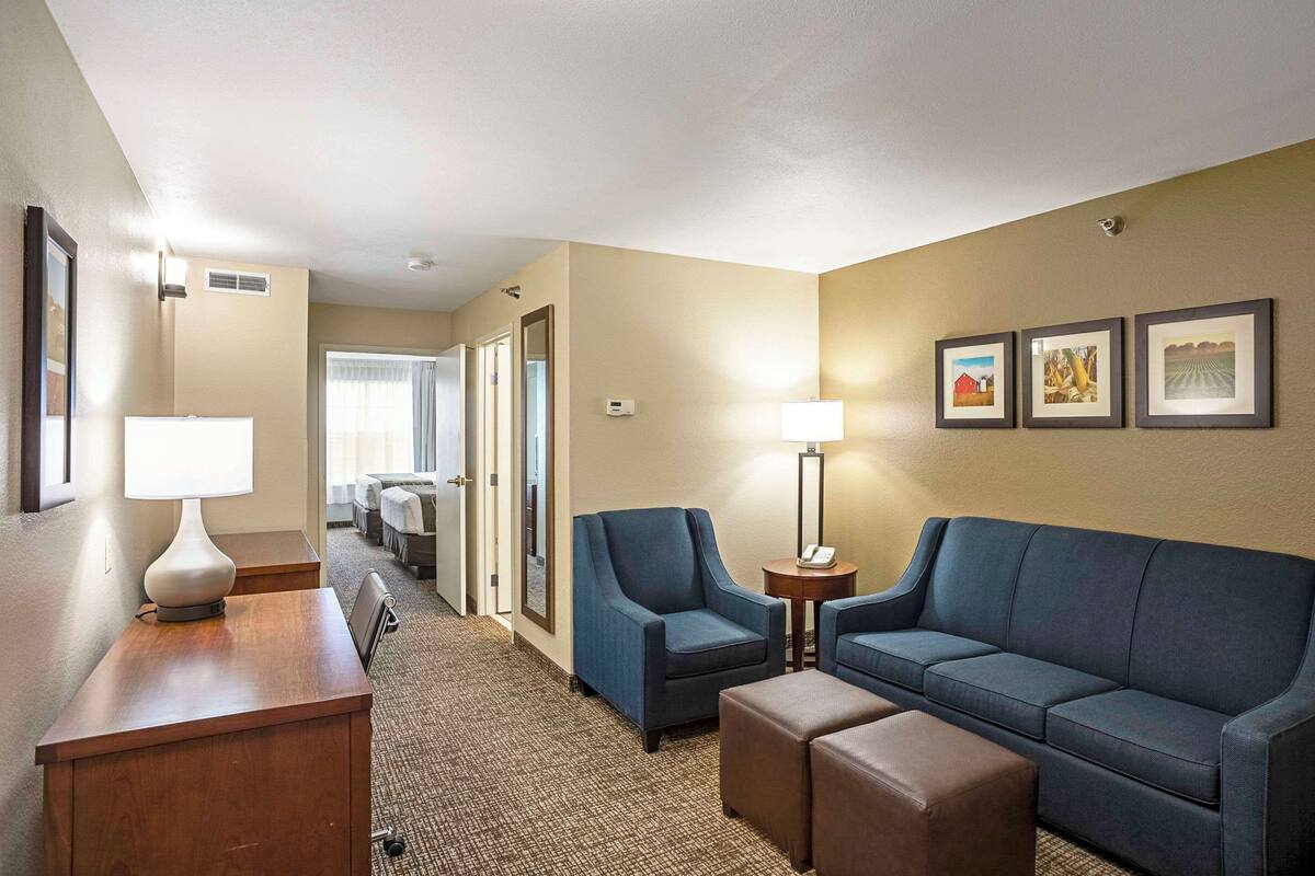 A hotel room with a wooden desk, two armchairs, a sofa, and a small table with a lamp. There are framed pictures on the wall and a carpeted floor.
