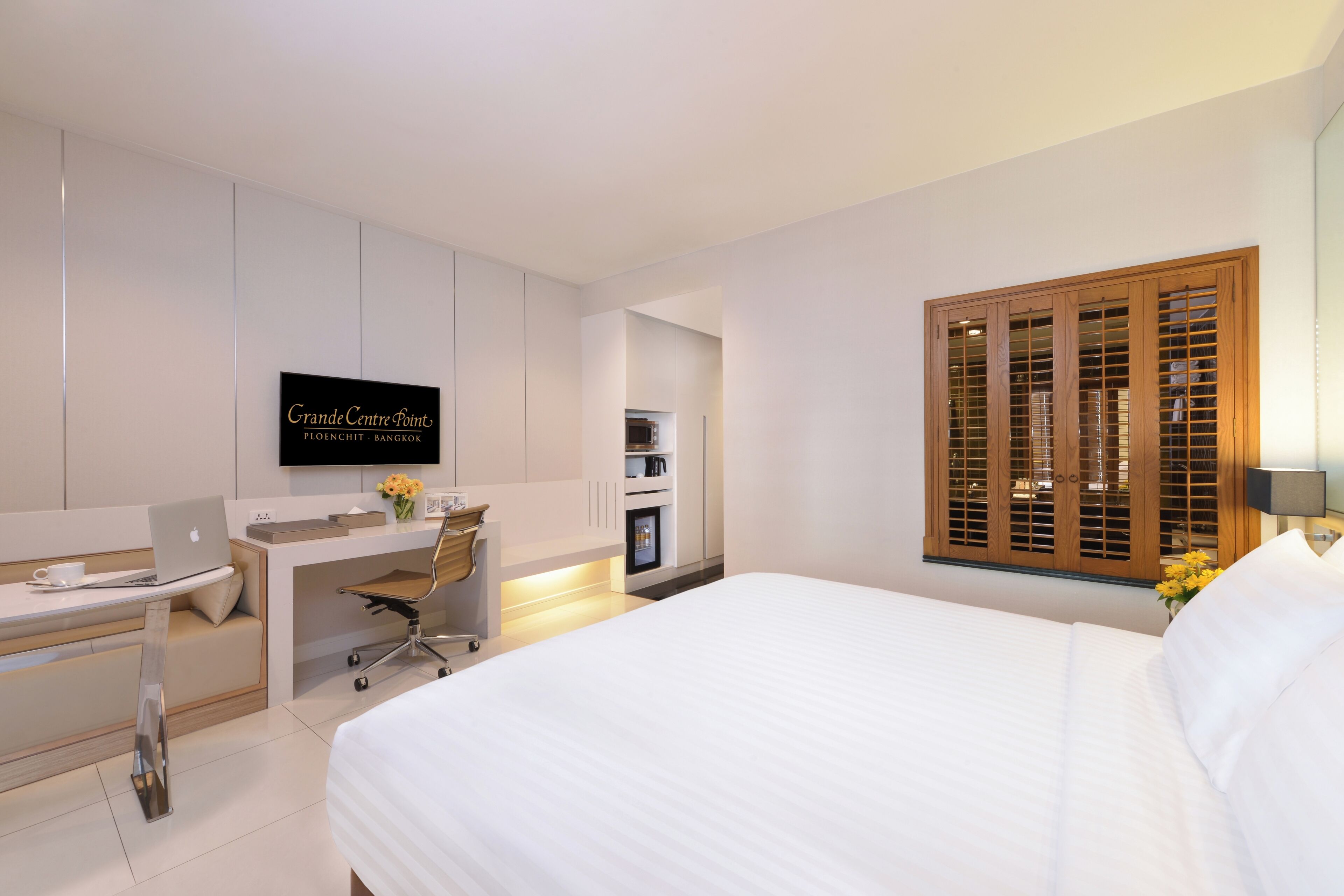 A modern hotel room with a bed, a desk with a laptop, a chair, a TV, and a window with wooden shutters.