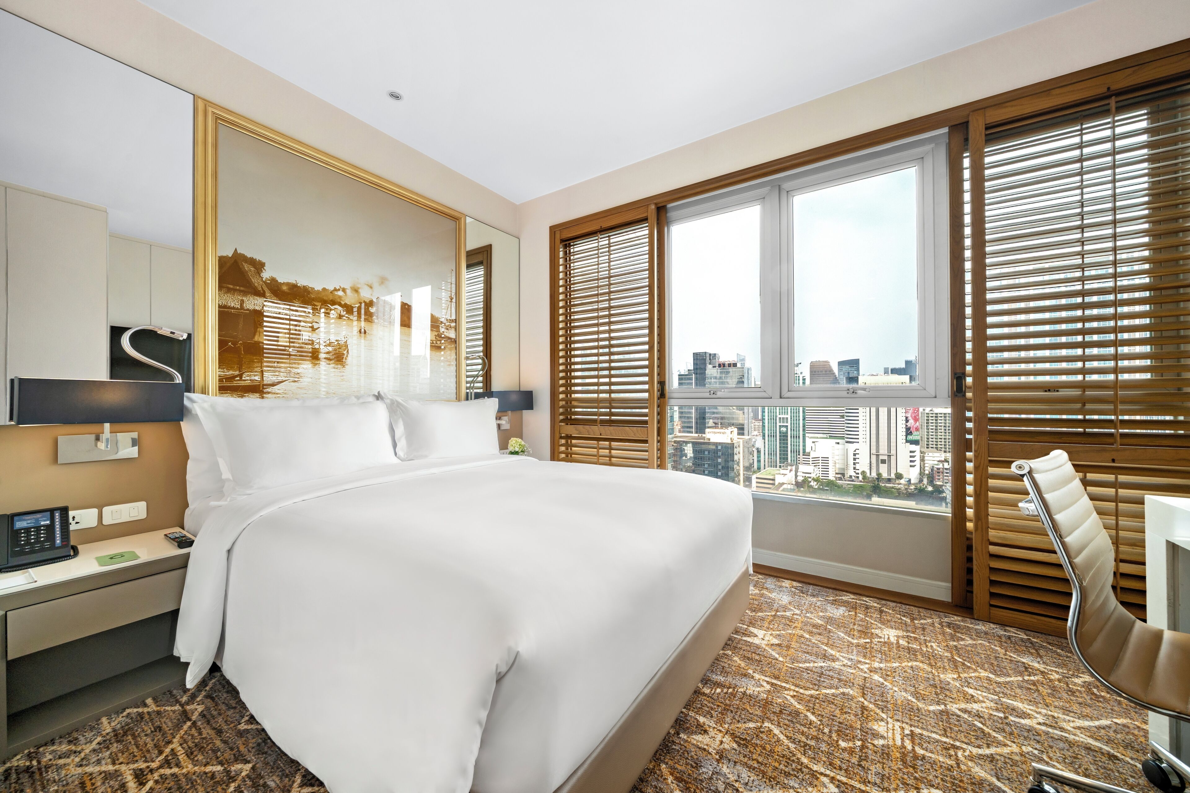 A hotel room with a large bed, a desk with a phone, a chair, a large window with blinds, and a view of the city.