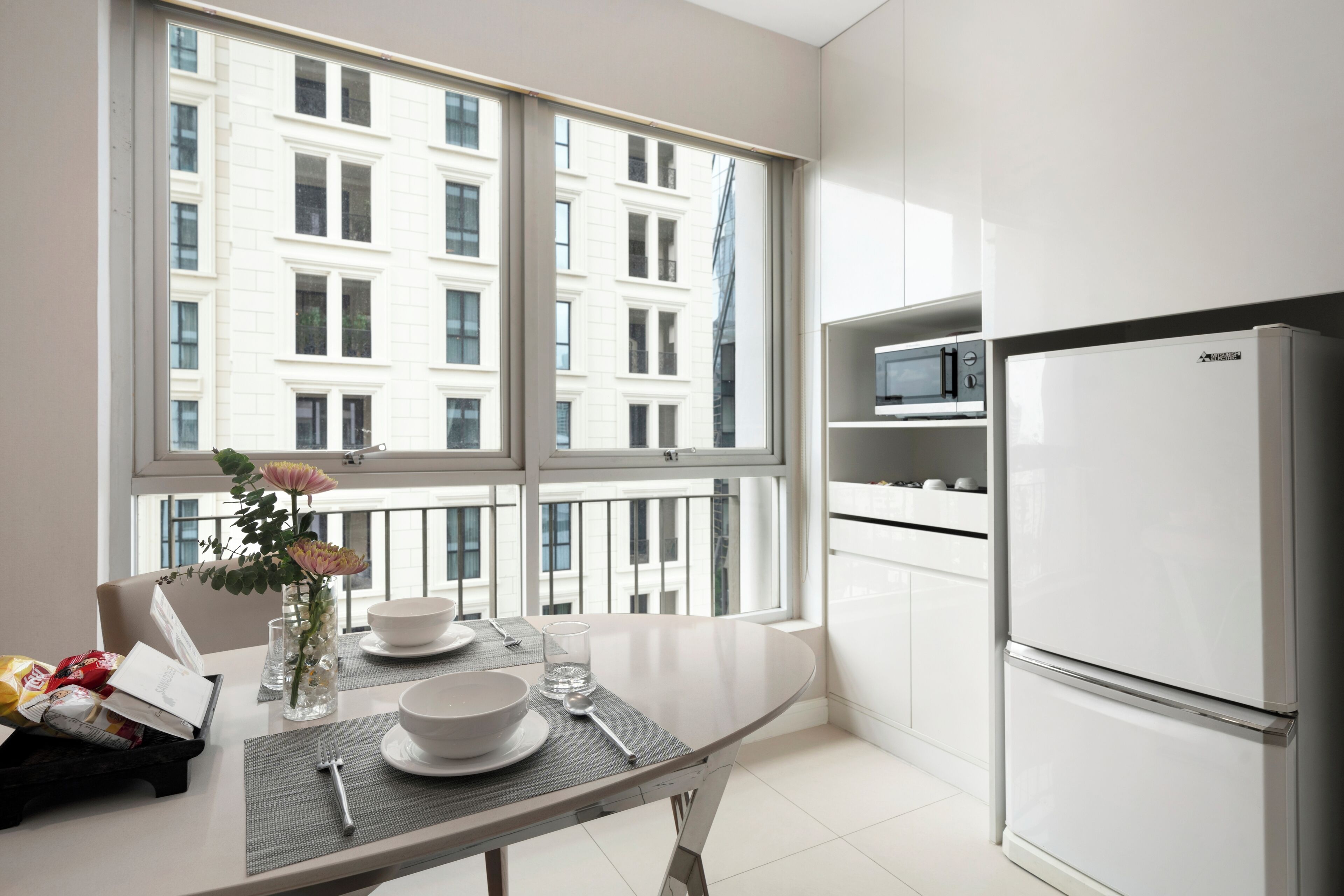A modern kitchen with a large window, white cabinets, a stainless steel refrigerator, and a dining table set with plates, glasses, and a vase with flowers.