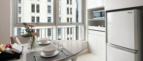 A modern kitchen with a large window, white cabinets, a stainless steel refrigerator, and a dining table set with plates, glasses, and a vase with flowers.