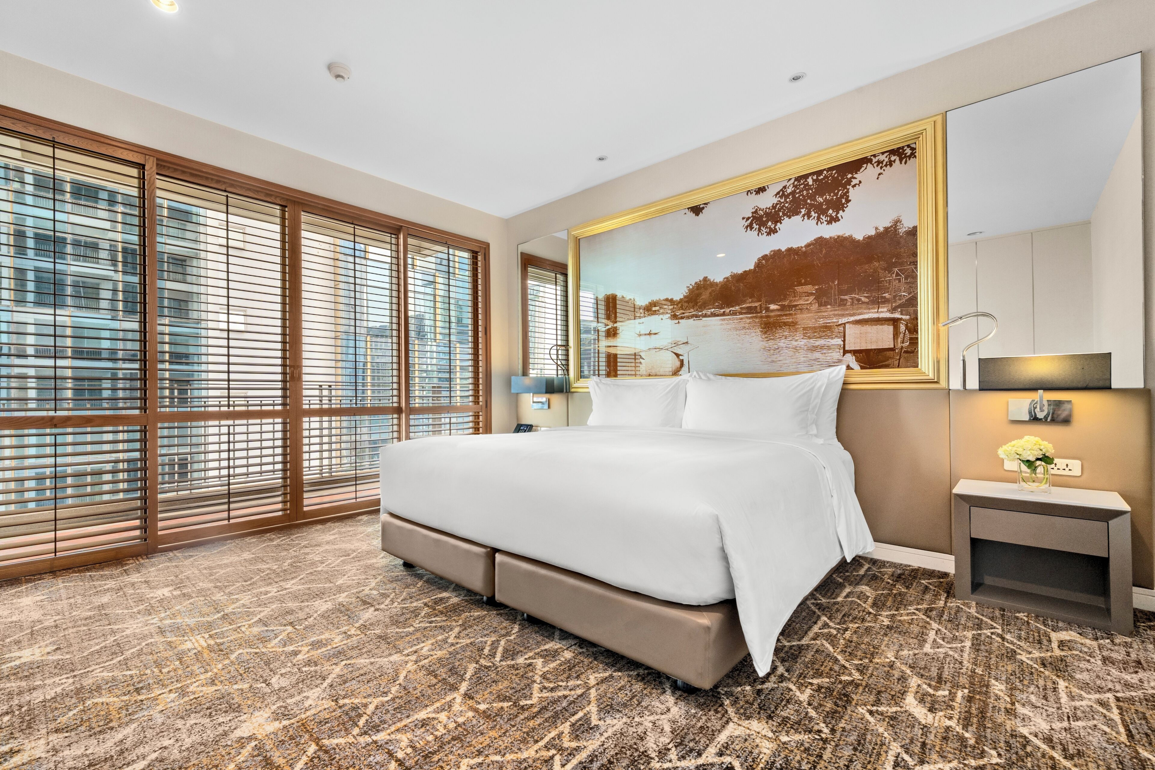 A hotel room with a large bed, a view of a cityscape through floor-to-ceiling windows, a large framed picture on the wall, and a small bedside table with a lamp.
