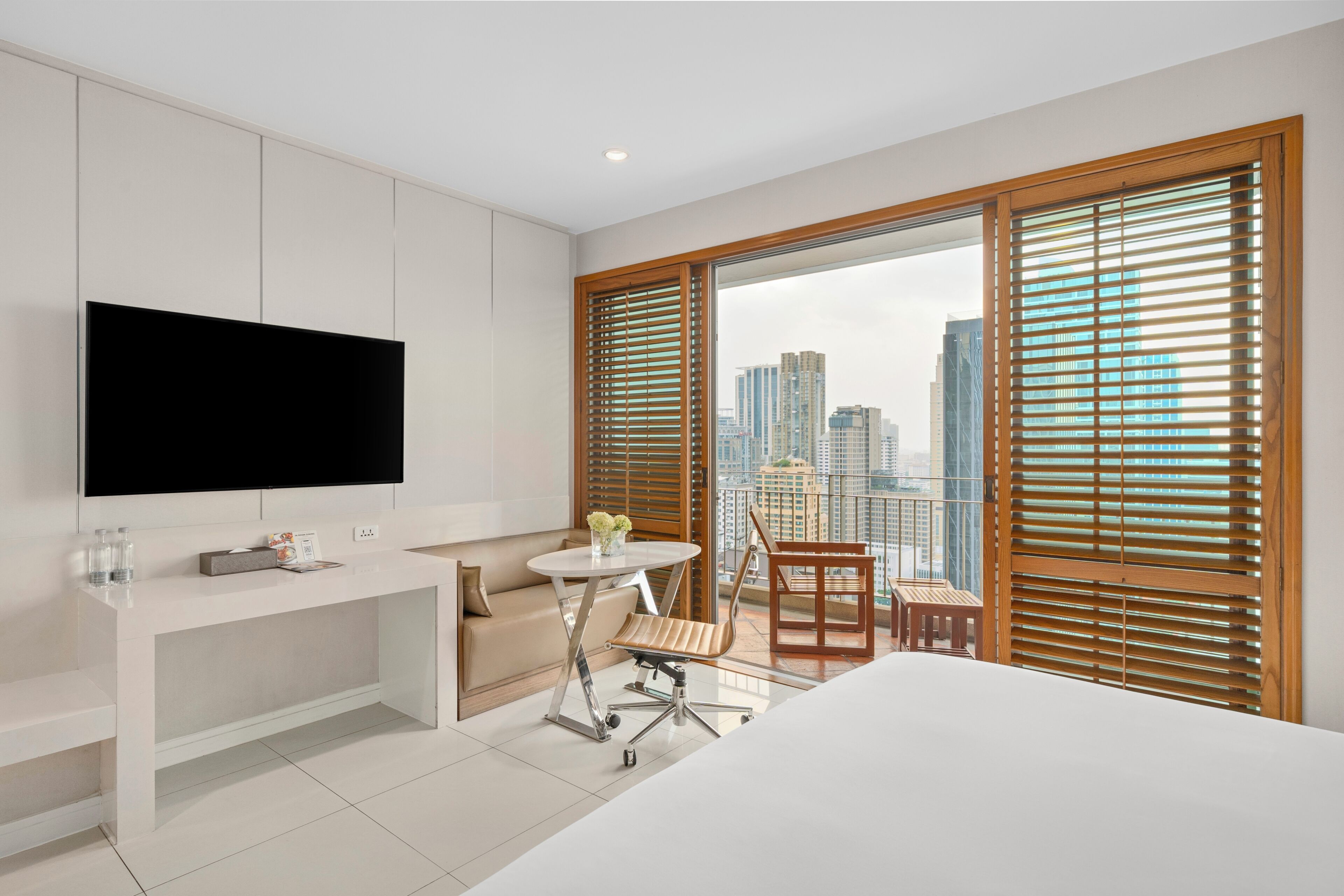 A modern hotel room with a large bed, a flat-screen TV, a small round table with a chair, a sofa, and a balcony with wooden blinds.