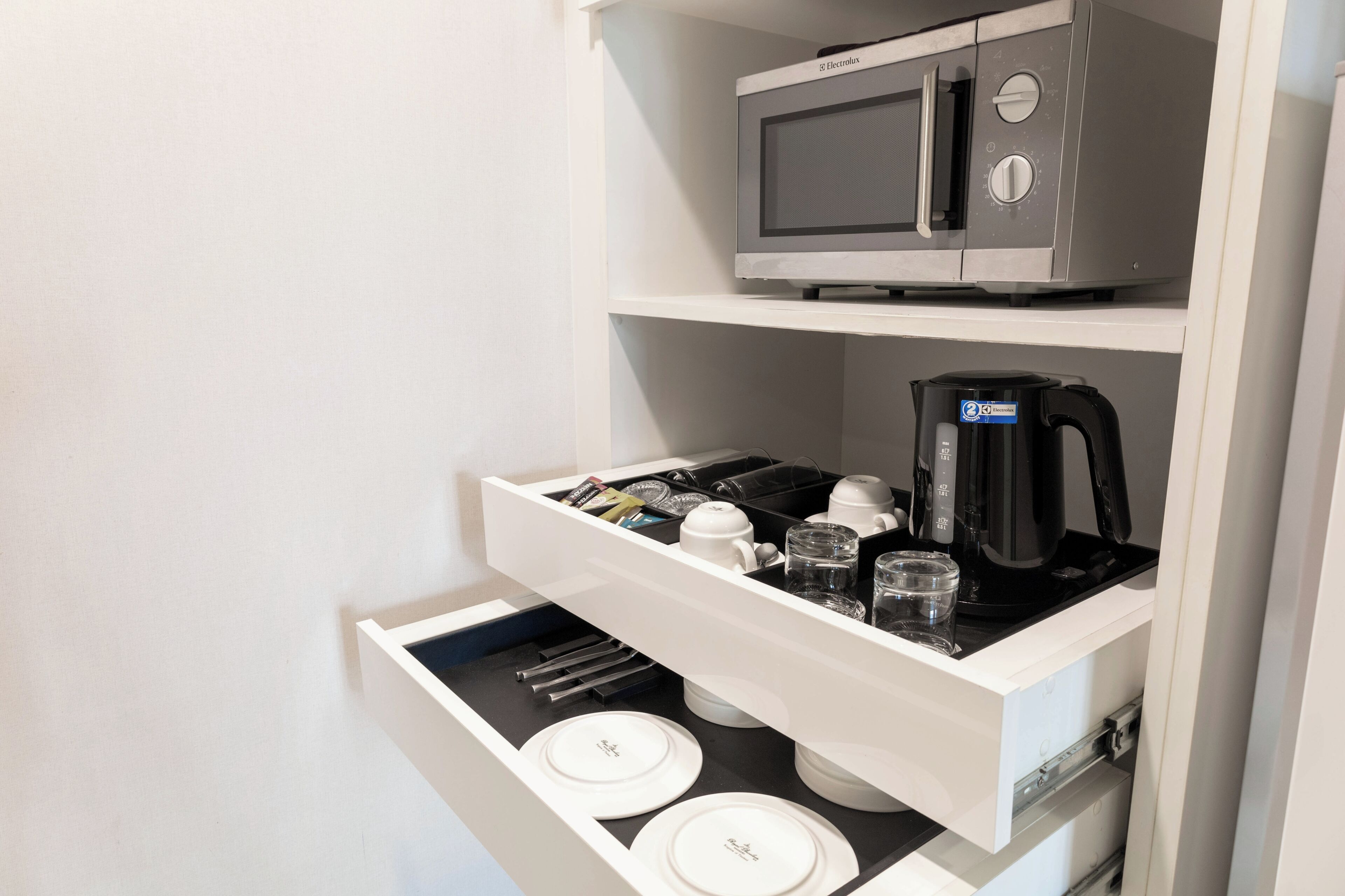A kitchen area with a microwave, coffee maker, and a drawer containing plates and utensils.