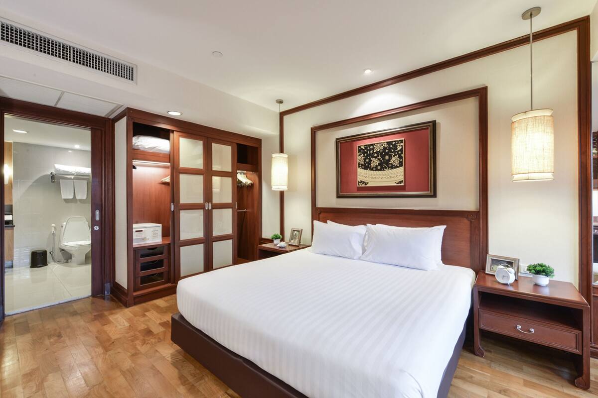 A hotel room with a large bed, wooden headboard, a framed artwork above the bed, a nightstand with a lamp, and a wooden wardrobe.