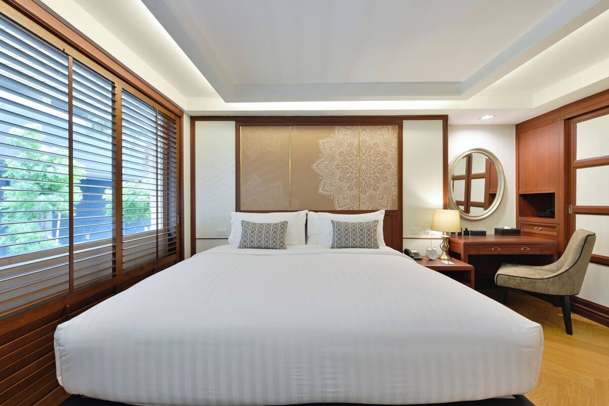 A large bed with a headboard, a bedside table with a lamp, a chair, and a desk with a mirror.