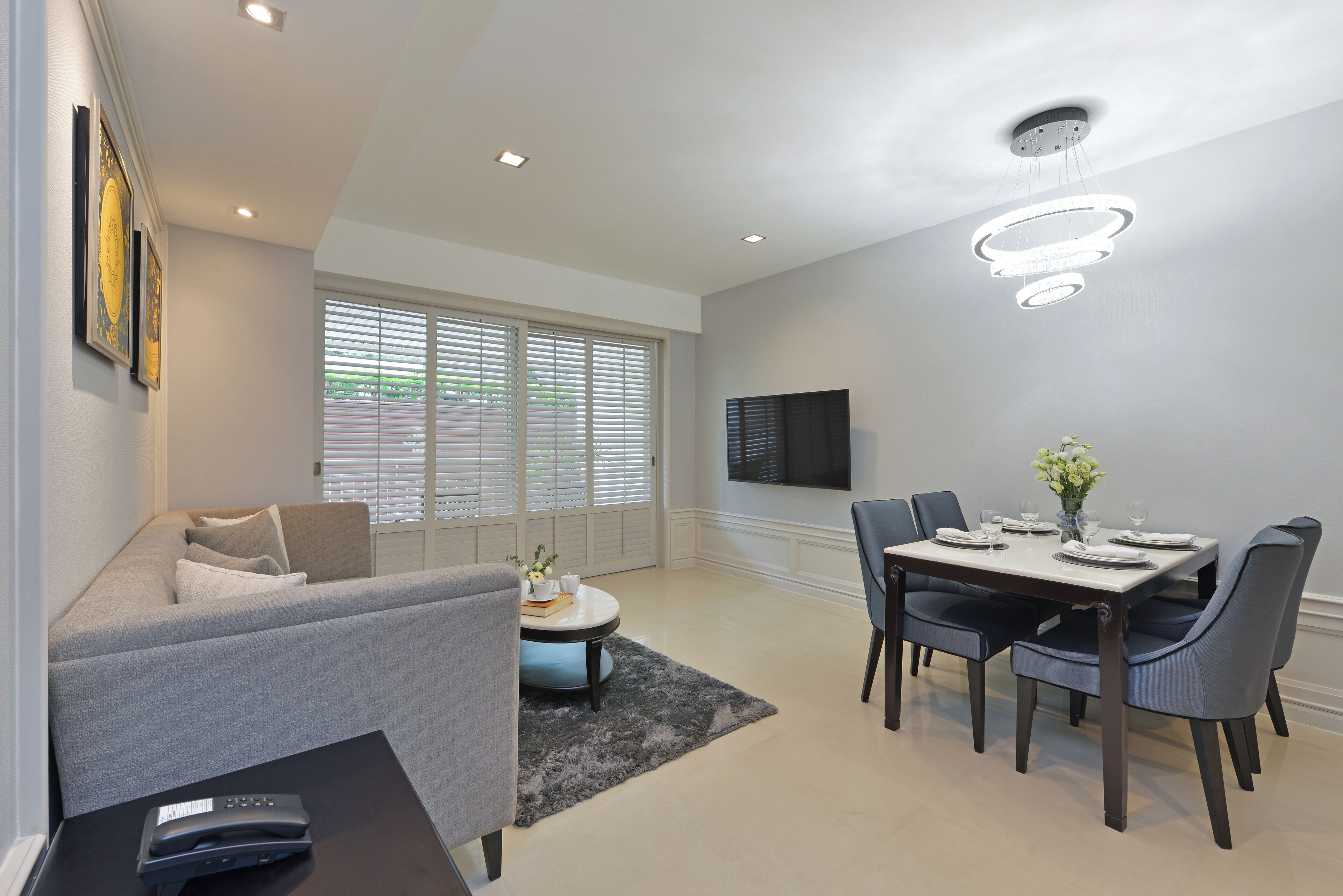 A modern living room with a grey sofa, a dining table set for four, and a flat-screen TV mounted on the wall.
