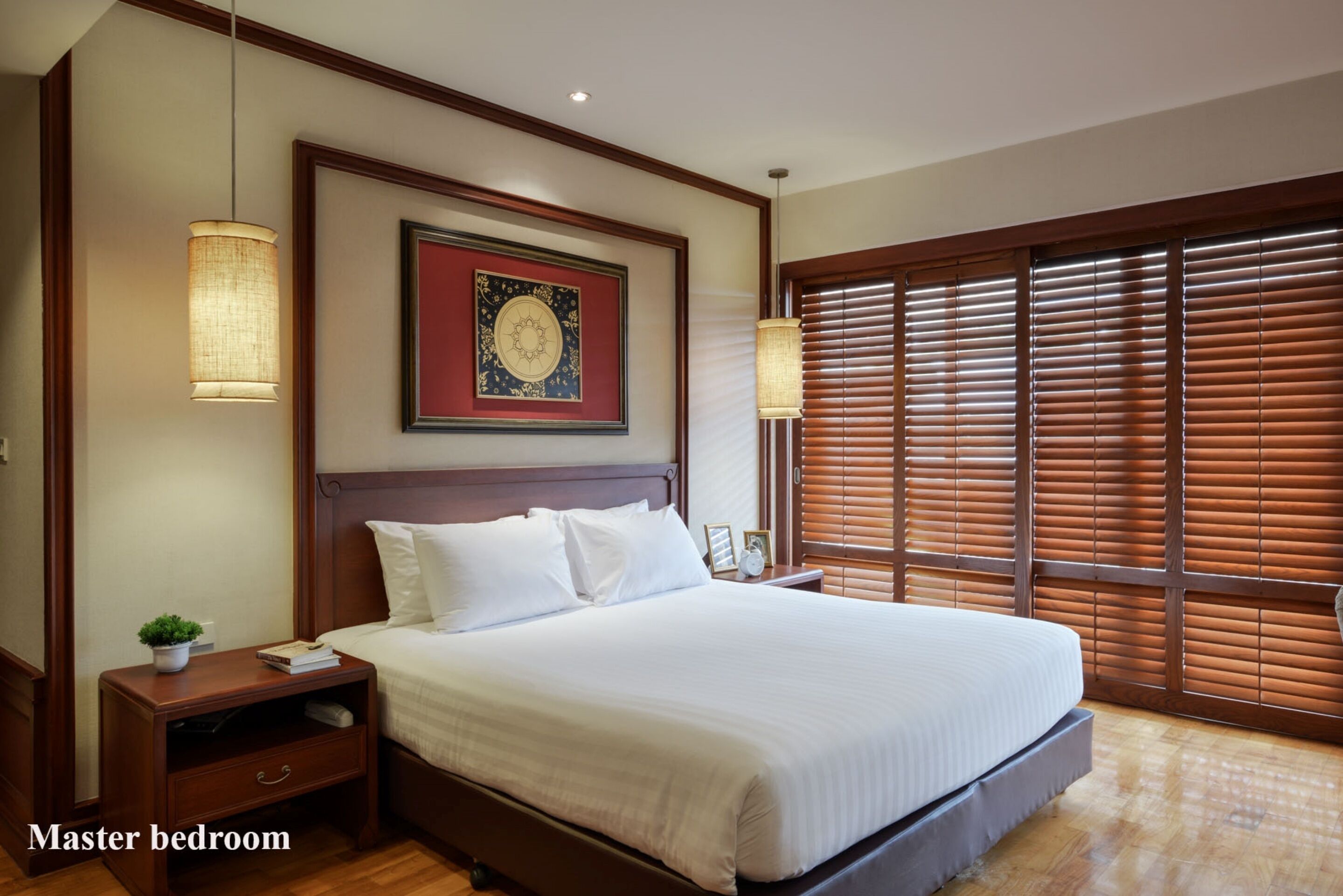 A master bedroom with a large bed, wooden headboard, two bedside tables, a nightstand, a wall-mounted lamp, a window with blinds, and a framed artwork above the bed.