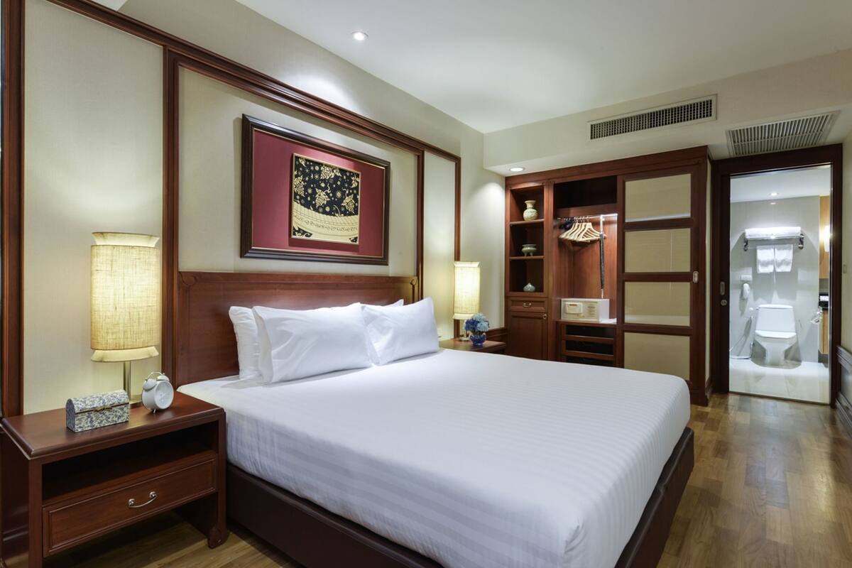 A hotel room with a large bed, wooden headboard, two bedside lamps, a nightstand, a framed picture on the wall, a walk-in closet, and a glass-enclosed bathroom.