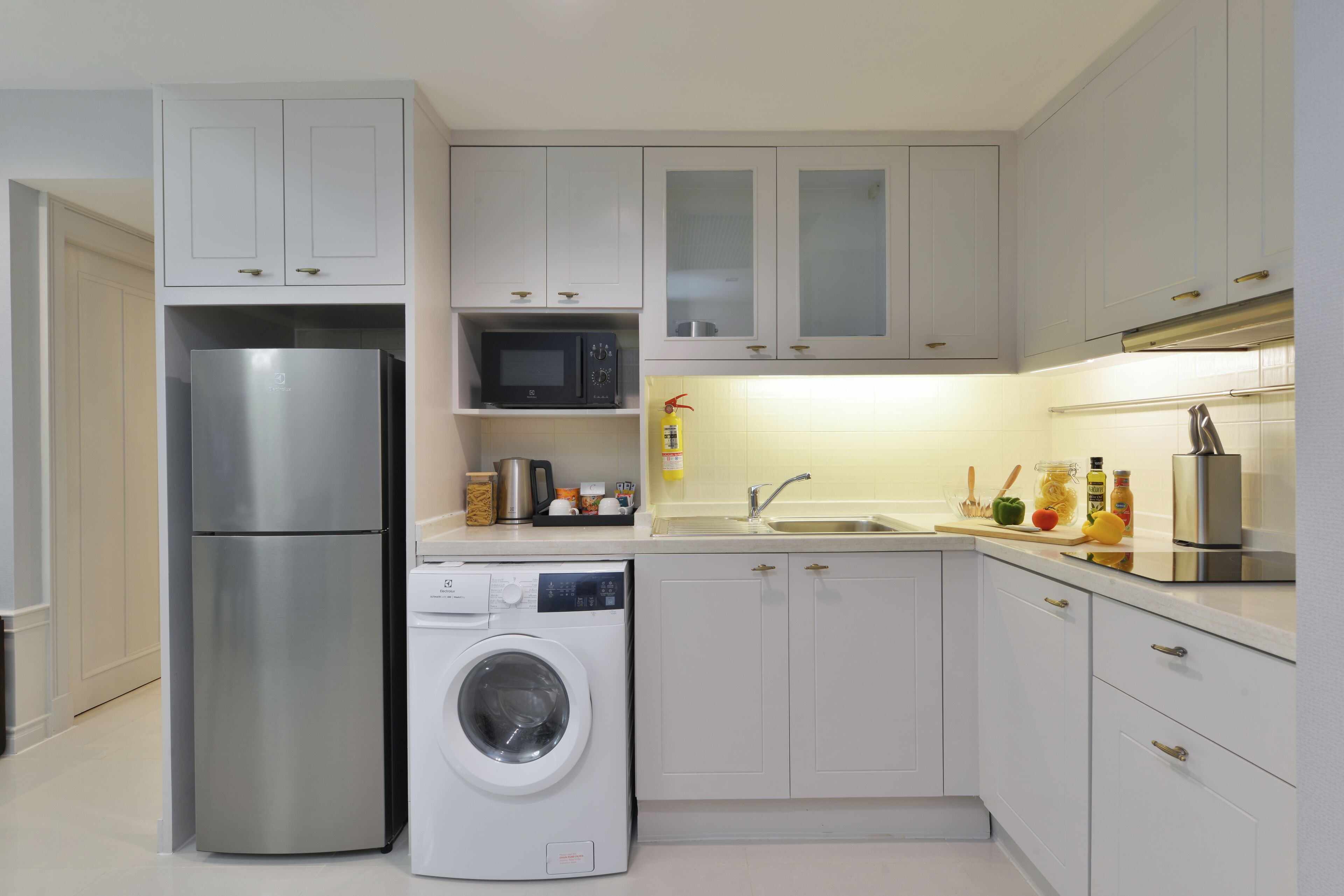 Eine moderne Küche mit weißen Schränken, einem Edelstahlkühlschrank, einer Waschmaschine, einer Mikrowelle und einem Waschbecken mit Armatur aus Edelstahl.