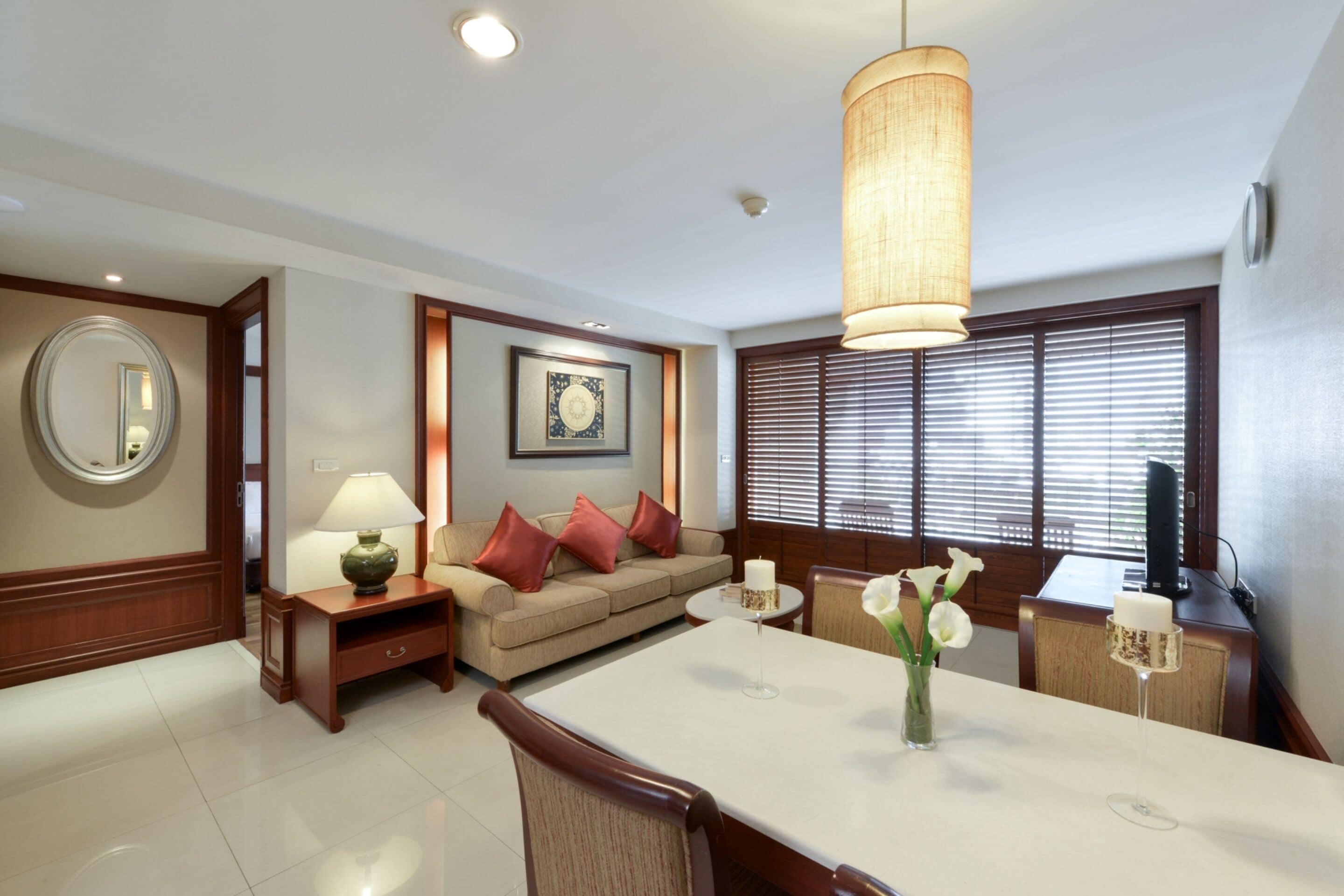 A modern living room with a dining area, a sofa with red cushions, a round mirror, and a large window with blinds.