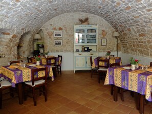 A rustic dining area with stone walls and a vaulted ceiling, featuring wooden tables and chairs, a central white cabinet with glass doors, and a tiled floor. - Montepaolo Dimora di Charme (Conversano)