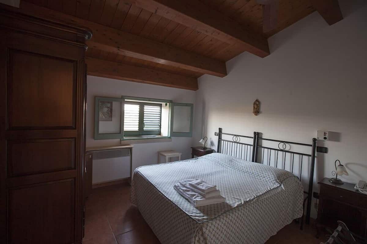 A bedroom with a wooden ceiling, a bed with a patterned bedspread, a nightstand with a lamp, and a small table with a telephone.