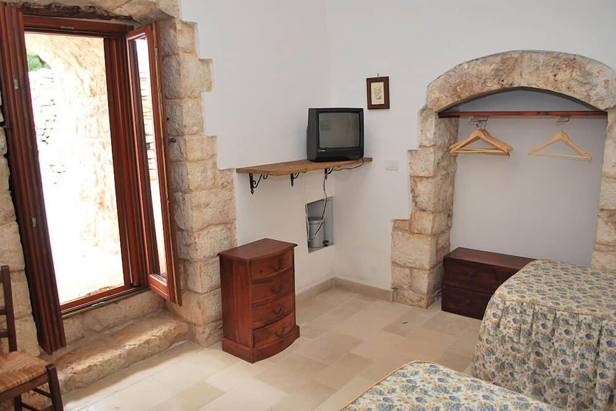 A room with stone walls, a wooden dresser, a television on a shelf, a bed with a floral bedspread, and a wooden chair.