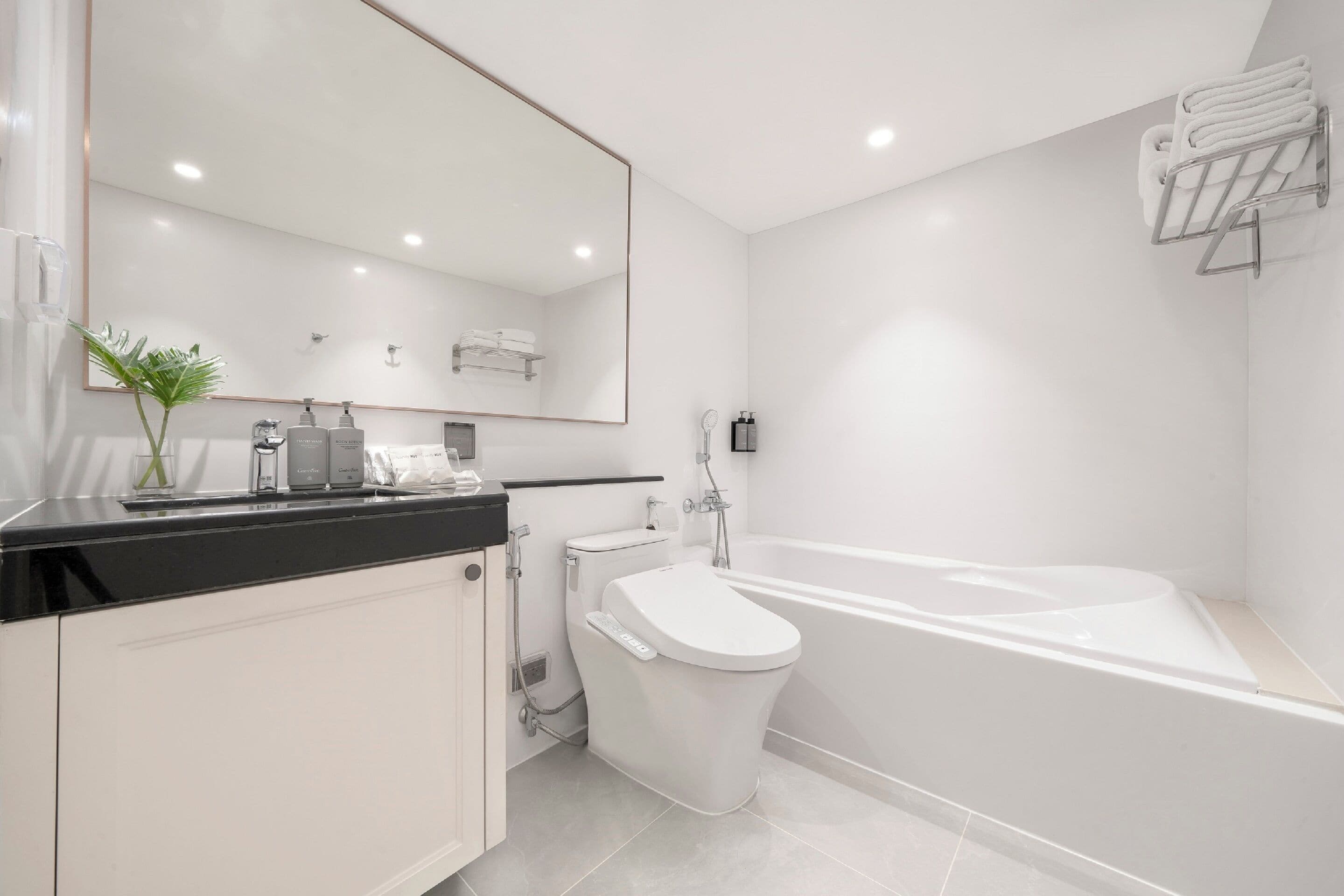 A modern bathroom with a white toilet, a bathtub, a large mirror, and a black countertop with toiletries.
