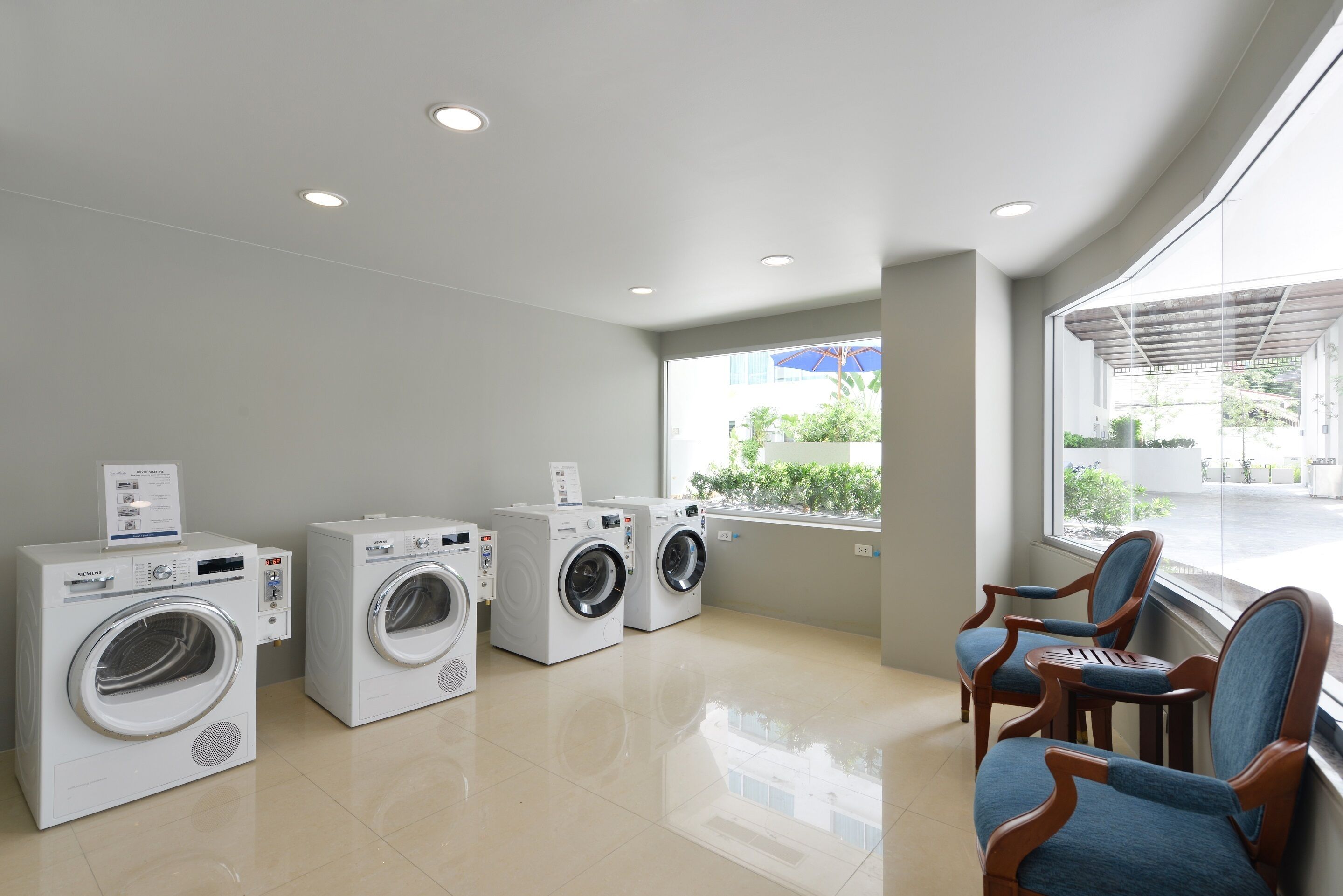 A laundry room with three white washing machines, wooden chairs, and a large window with a view of greenery.
